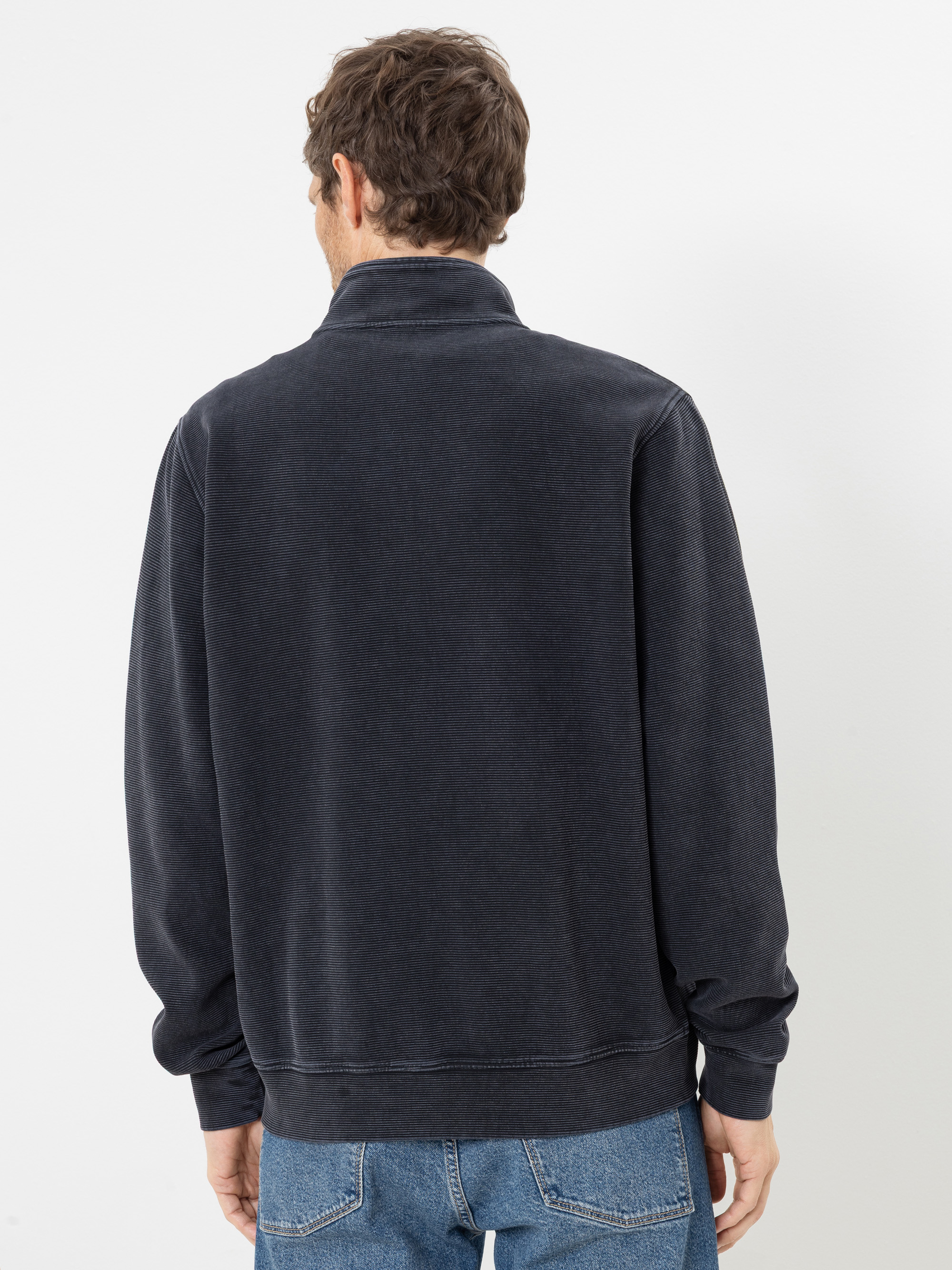 Vor einem schlichten weißen Hintergrund ist eine Person mit kurzen braunen Haaren von hinten zu sehen, die eine ausgewaschene schwarze Jacke mit Reißverschluss und blaue Jeans trägt.