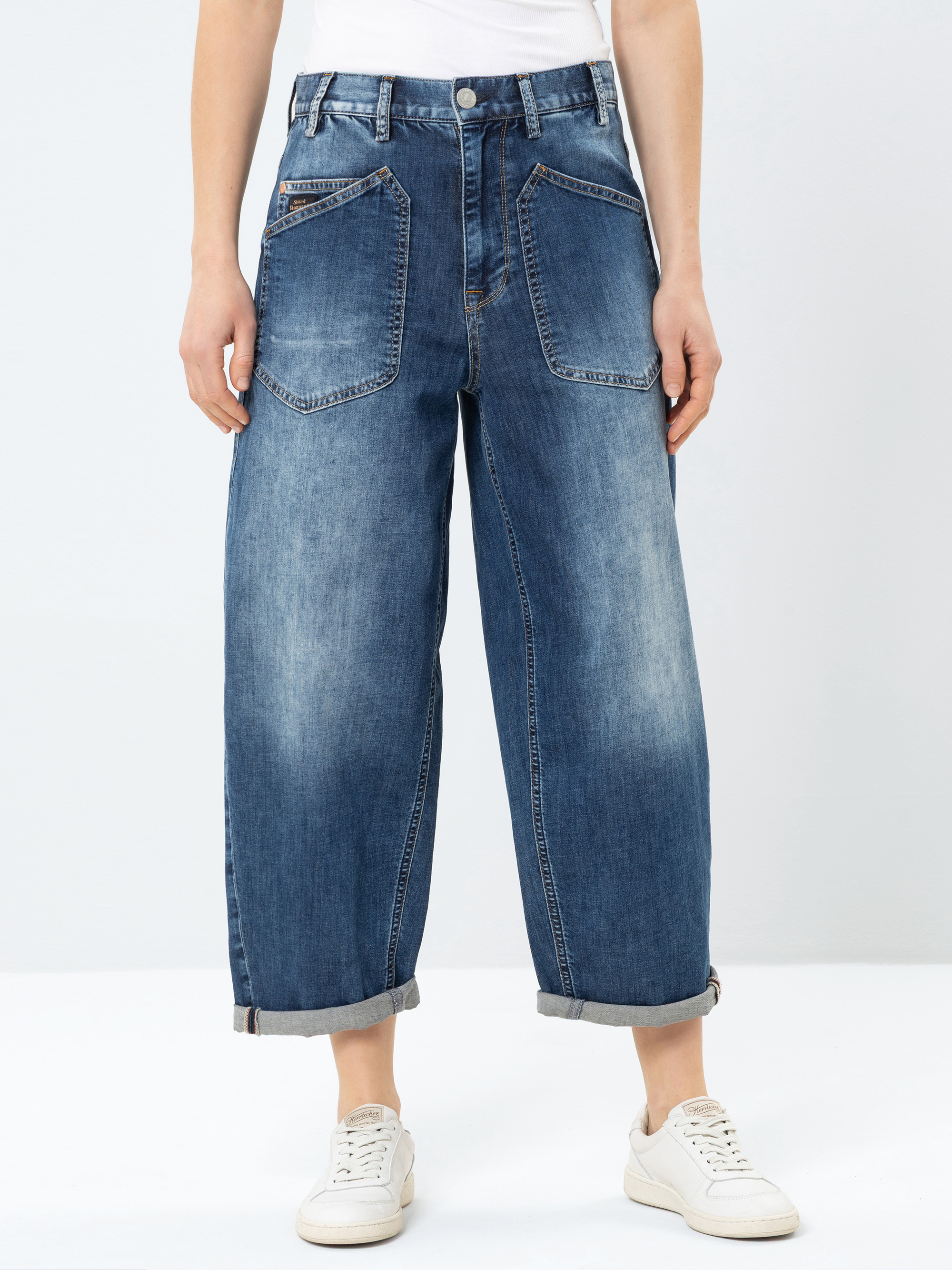 Eine Person, die ein weißes Hemd, eine locker sitzende blaue Jeans mit großen Vordertaschen und aufgerollten Manschetten sowie weiße Turnschuhe trägt, steht vor einem schlichten hellen Hintergrund.