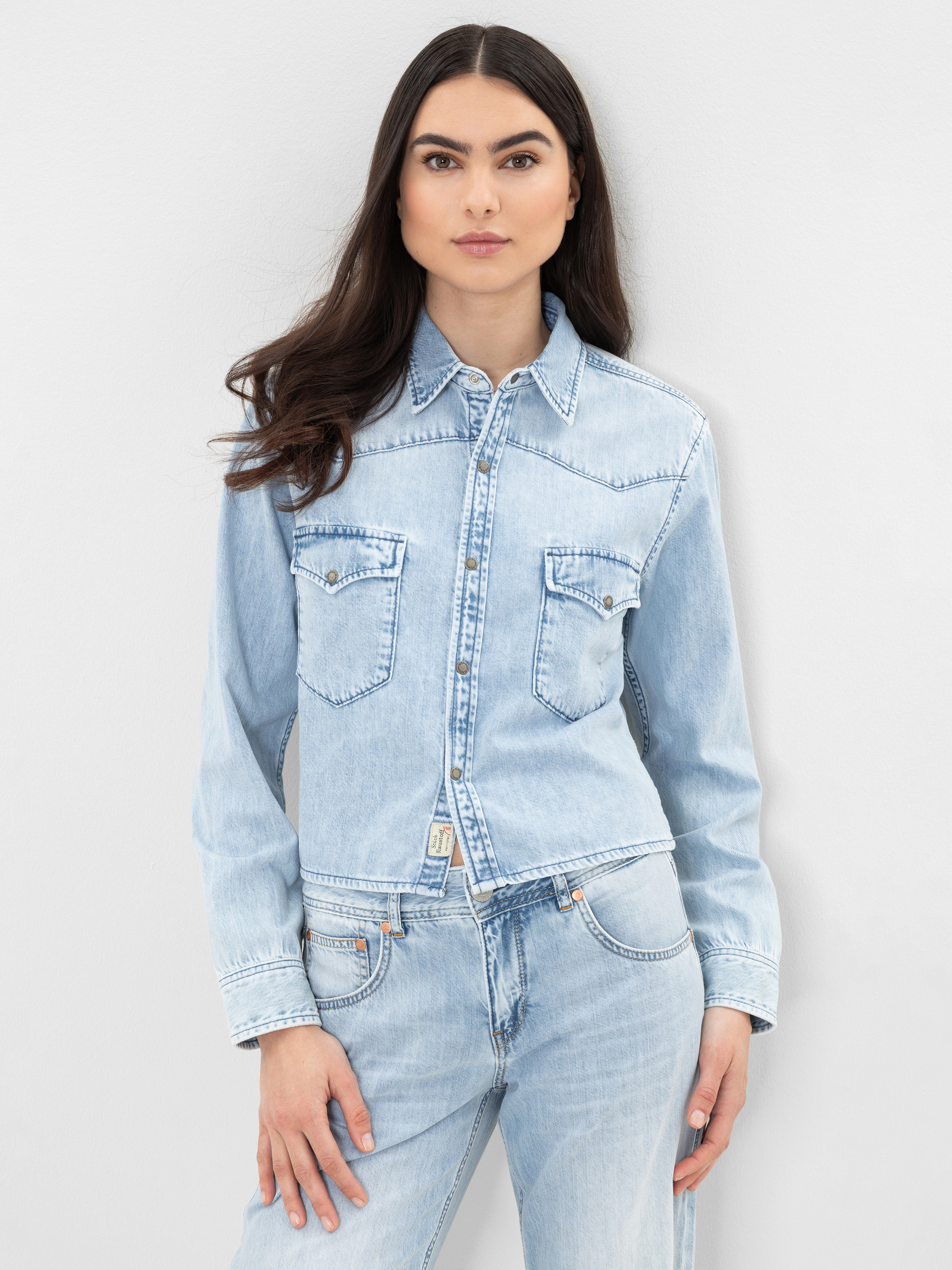Eine Frau mit langen dunklen Haaren trägt ein hellblaues Jeanshemd mit Knopfleiste und Brusttaschen und dazu passende hellblaue Jeans, die vor einem schlichten hellgrauen Hintergrund stehen.
