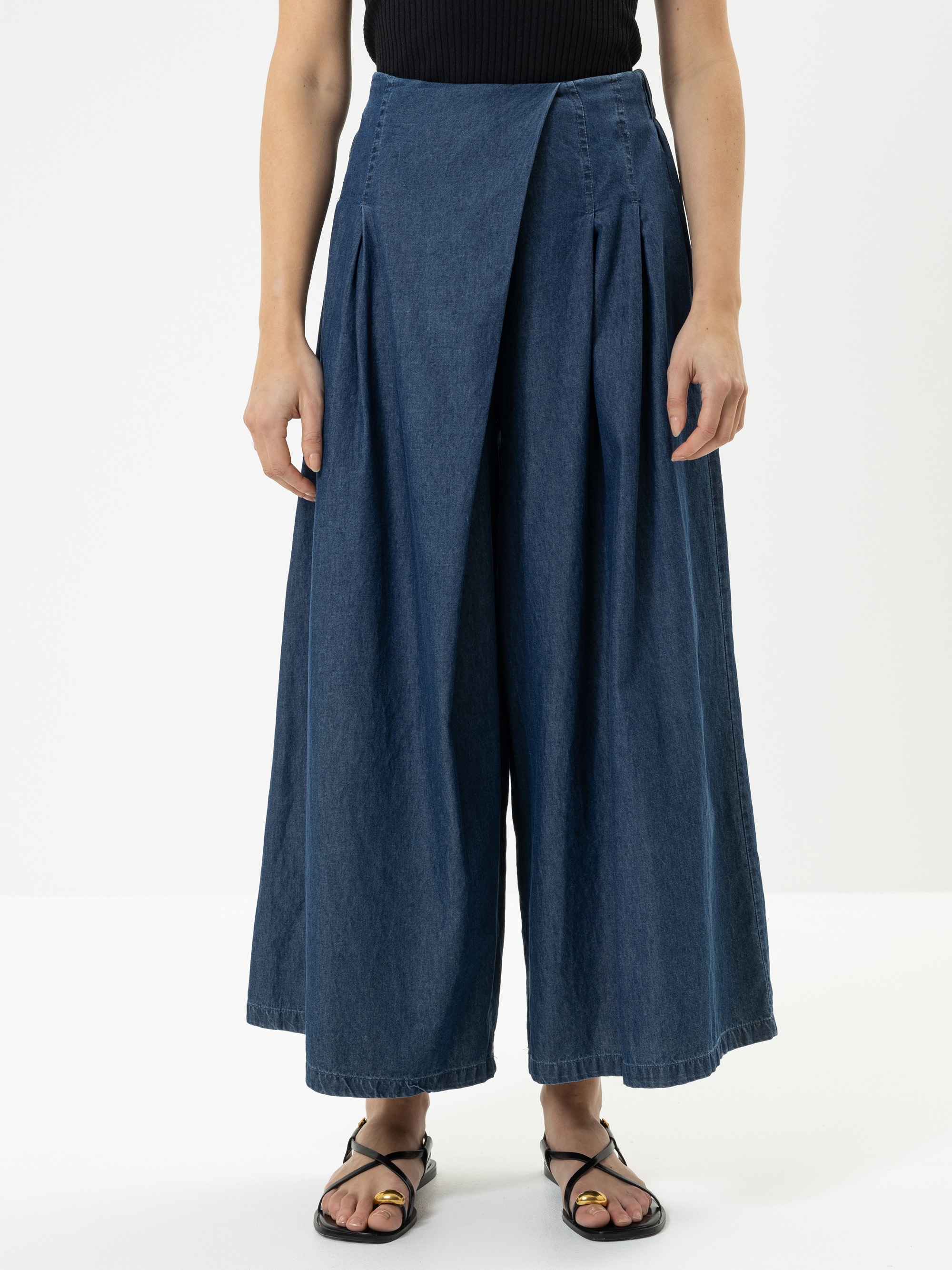 Eine Person trägt eine blaue Jeanshose mit weitem Bein und hoher Taille, die vorne in Falten gelegt ist, ein schwarzes ärmelloses Oberteil und schwarze Sandalen, die vor einem schlichten weißen Hintergrund stehen.