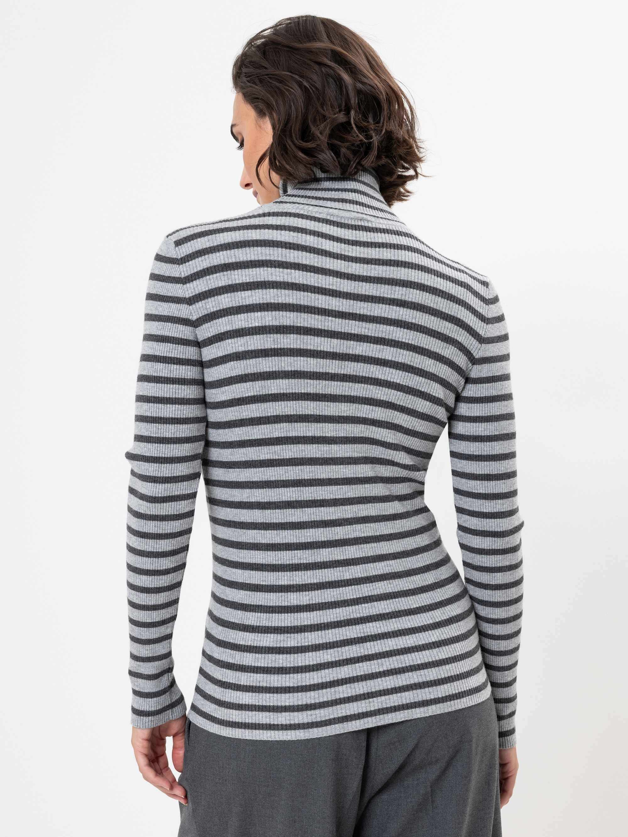 Eine Person mit kurzen braunen Haaren wird von hinten gezeigt, die einen grau-schwarz gestreiften langärmeligen Rollkragenpullover und eine graue Hose trägt und vor einem schlichten weißen Hintergrund steht.