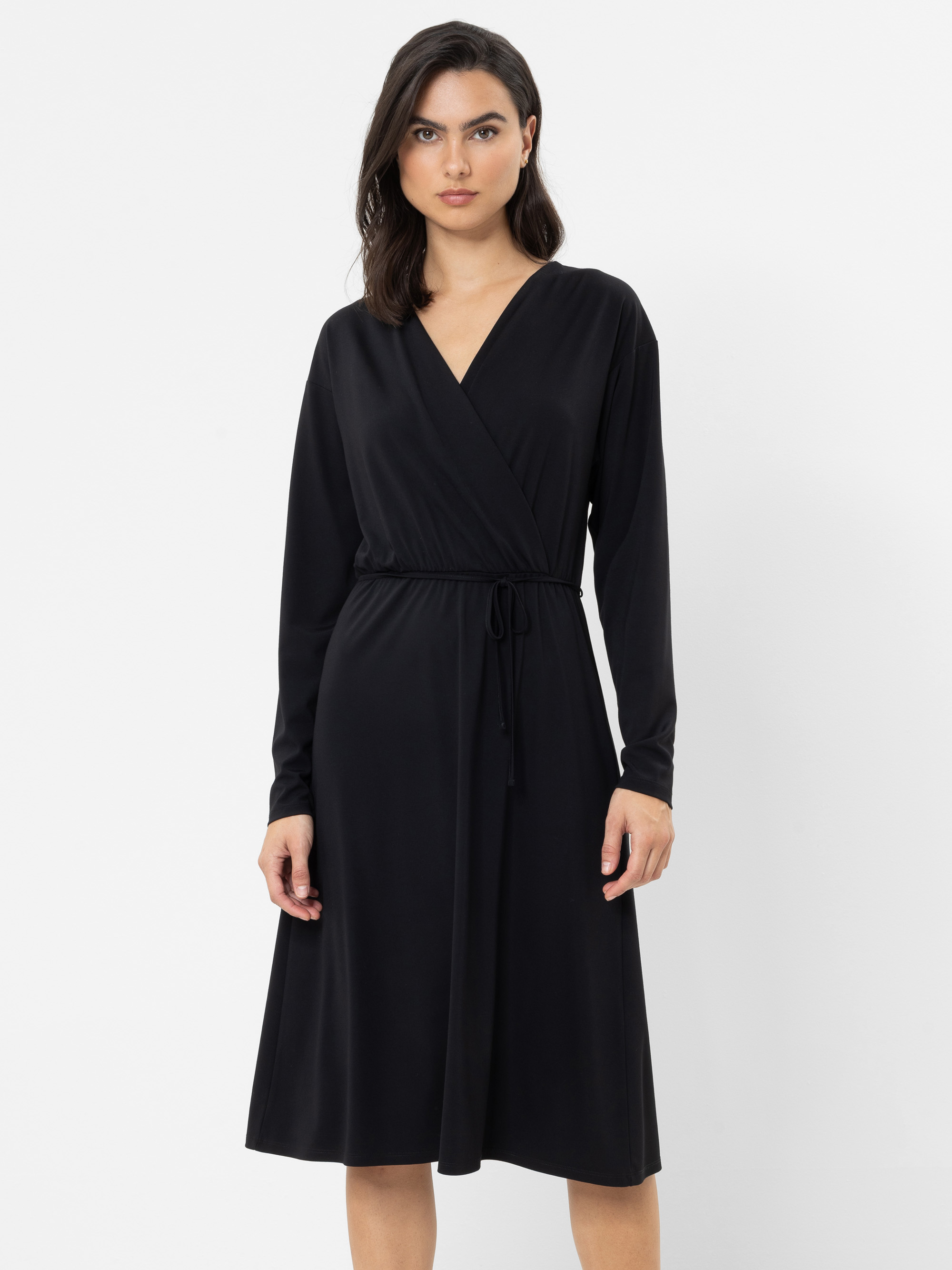 Vor einem schlichten weißen Hintergrund steht eine Frau, die ein schwarzes, langärmeliges, knielanges Wickelkleid mit einem Bindegürtel an der Taille trägt.