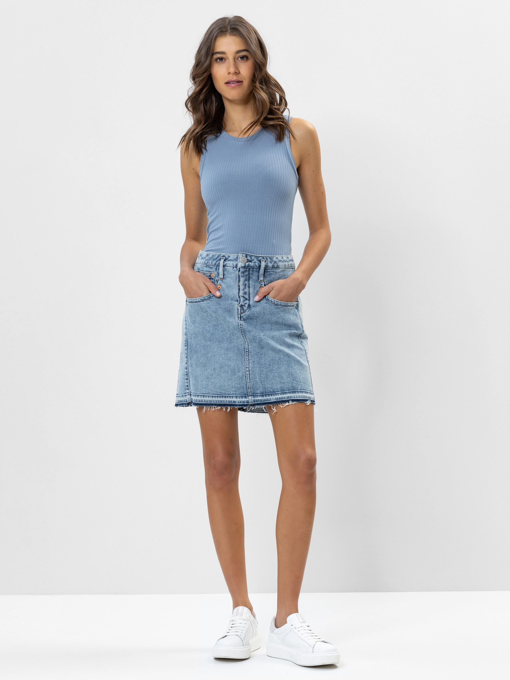 Eine Frau steht vor einem schlichten weißen Hintergrund und trägt ein ärmelloses blaues geripptes Oberteil, einen Jeansrock mit ausgefranstem Saum und weiße Turnschuhe. Ihre Hände stecken in den Rocktaschen, und sie hat gewelltes braunes Haar.