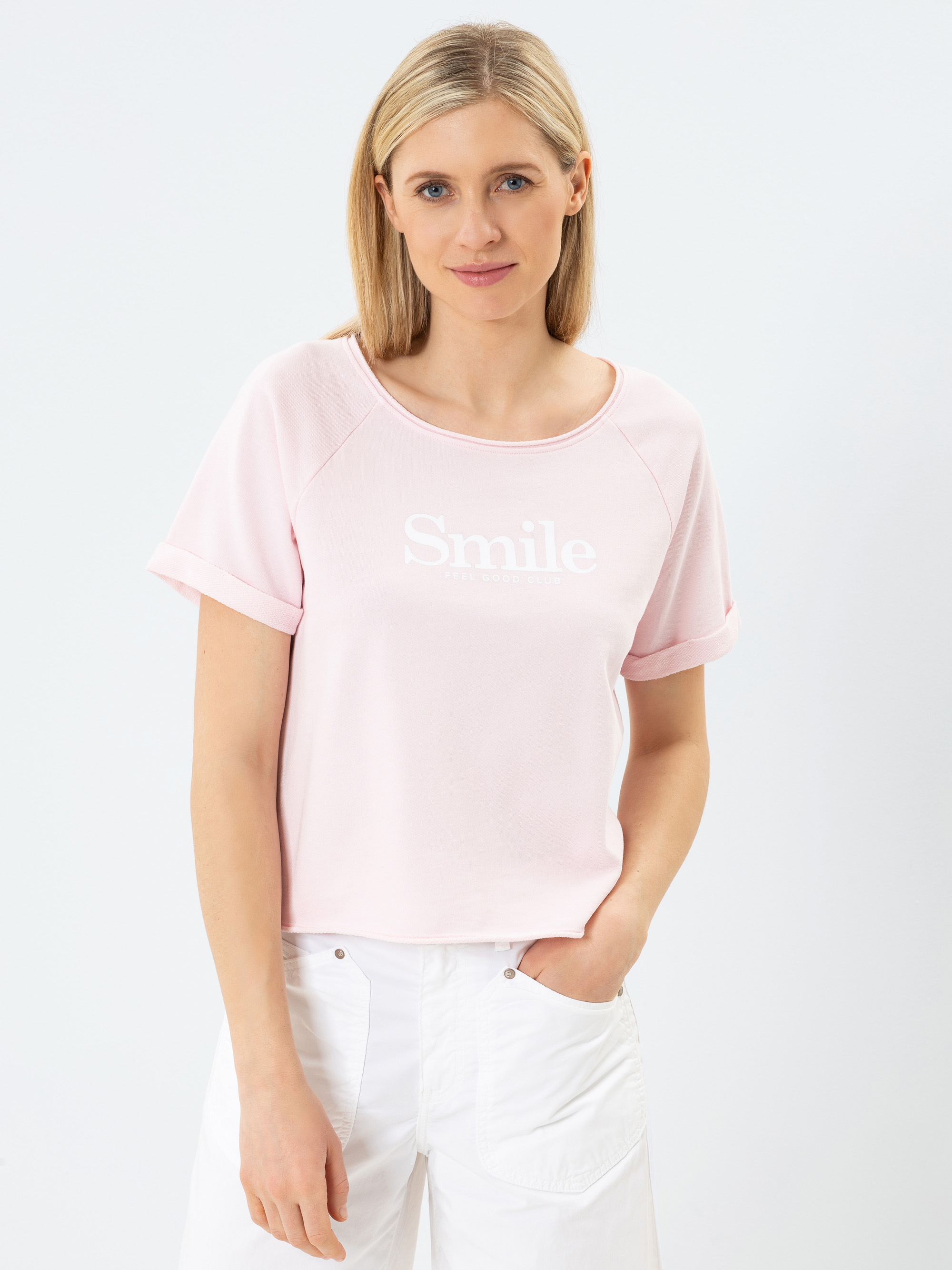 Eine Frau mit glattem blondem Haar, die ein hellrosa T-Shirt mit der Aufschrift Smile und eine weiße Hose trägt, steht mit einer Hand in der Tasche vor einem schlichten weißen Hintergrund.