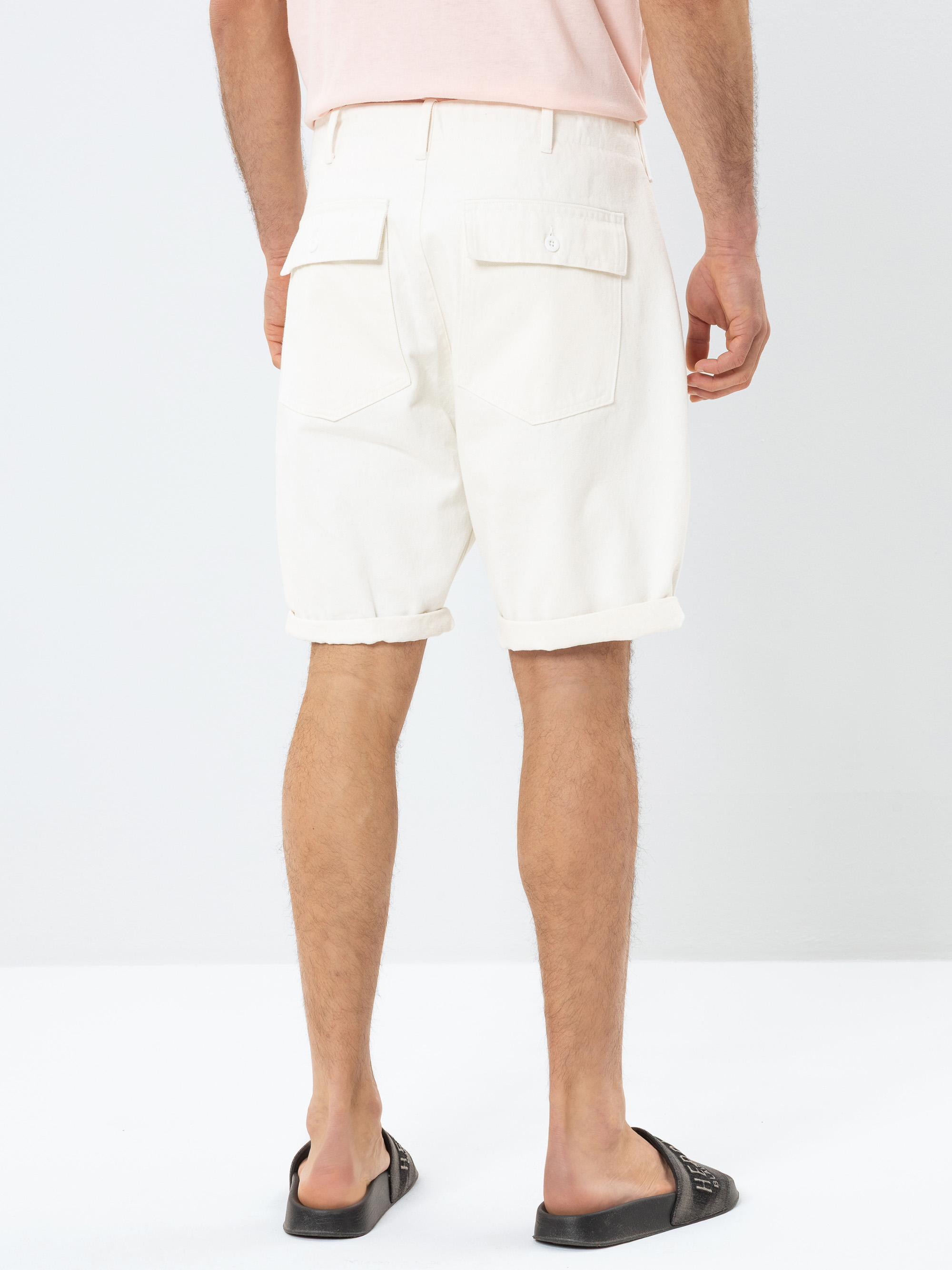 Ein Mann steht mit dem Gesicht zur Wand und trägt weiße Shorts mit Bündchen und Gesäßtaschen, ein hellrosa Hemd und schwarze Sandalen vor einem schlichten weißen Hintergrund.
