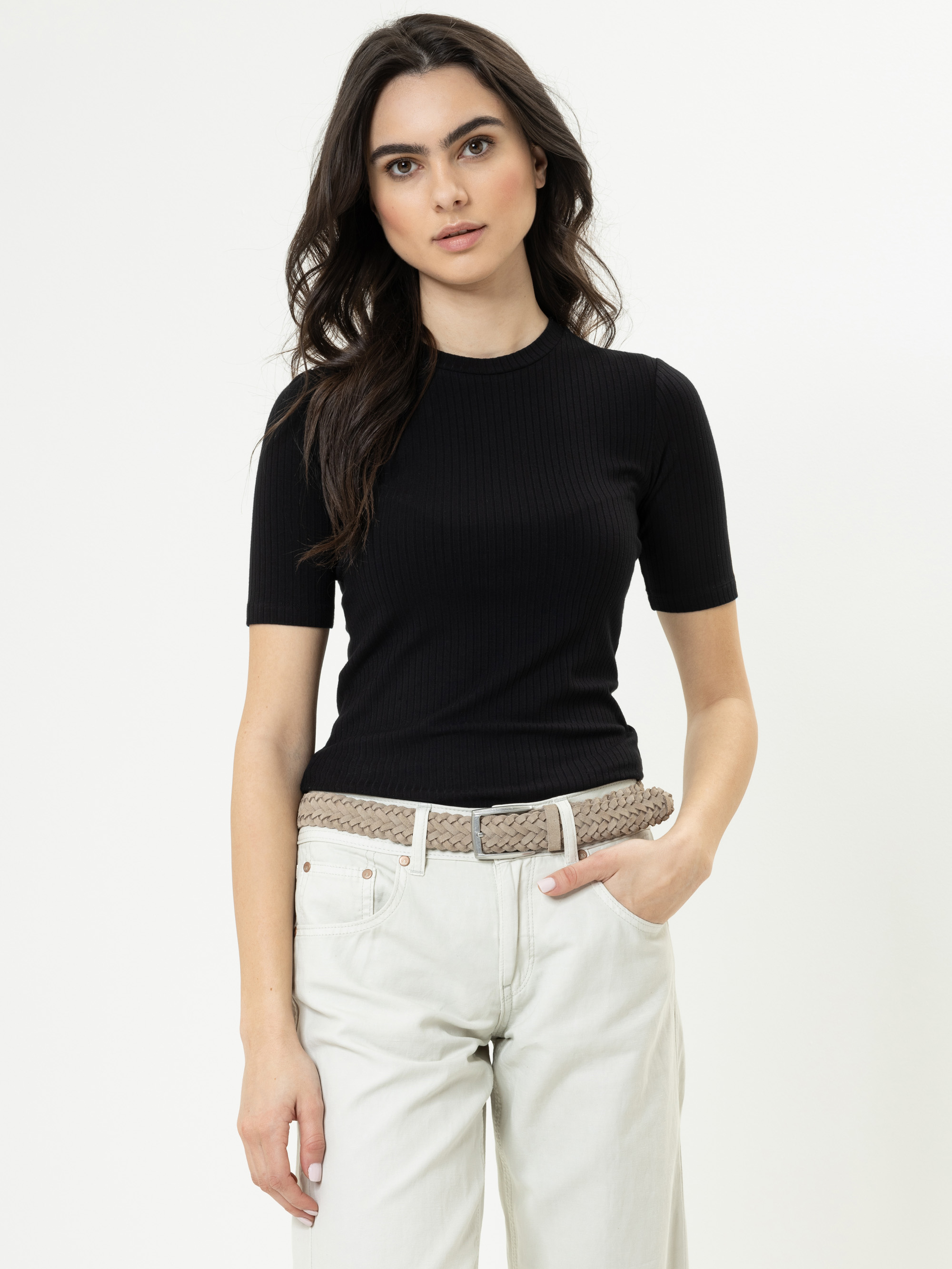 Eine Frau mit langen dunklen Haaren, die ein schwarzes, kurzärmeliges Oberteil, helle Jeans und einen gewebten beigen Gürtel trägt, steht vor einem schlichten weißen Hintergrund und schaut mit neutralem Blick in die Kamera.