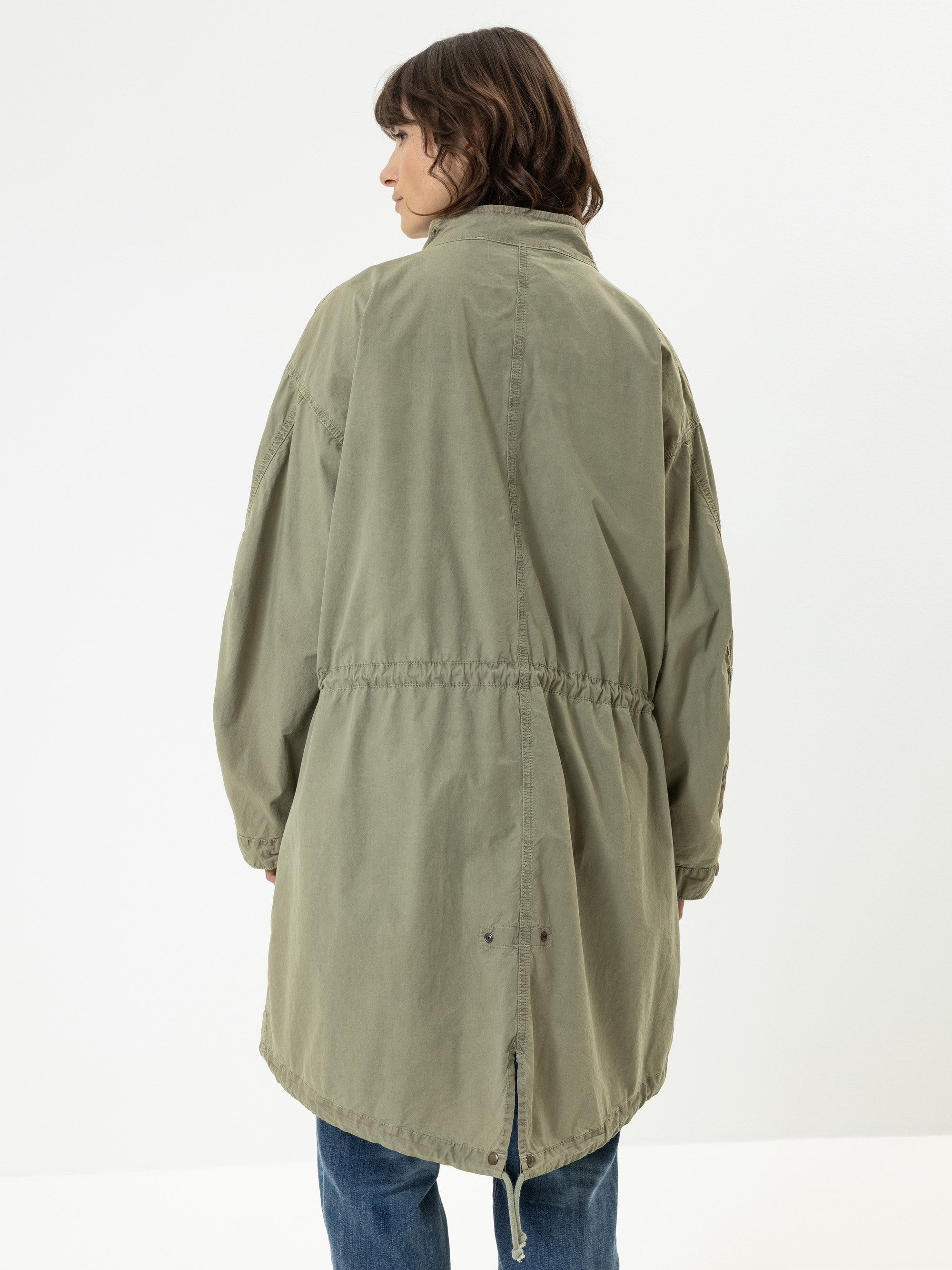 Eine Person mit schulterlangen braunen Haaren, die eine übergroße olivgrüne Jacke mit Kordelzug in der Taille trägt, steht mit dem Rücken zur Kamera vor einem schlichten weißen Hintergrund.