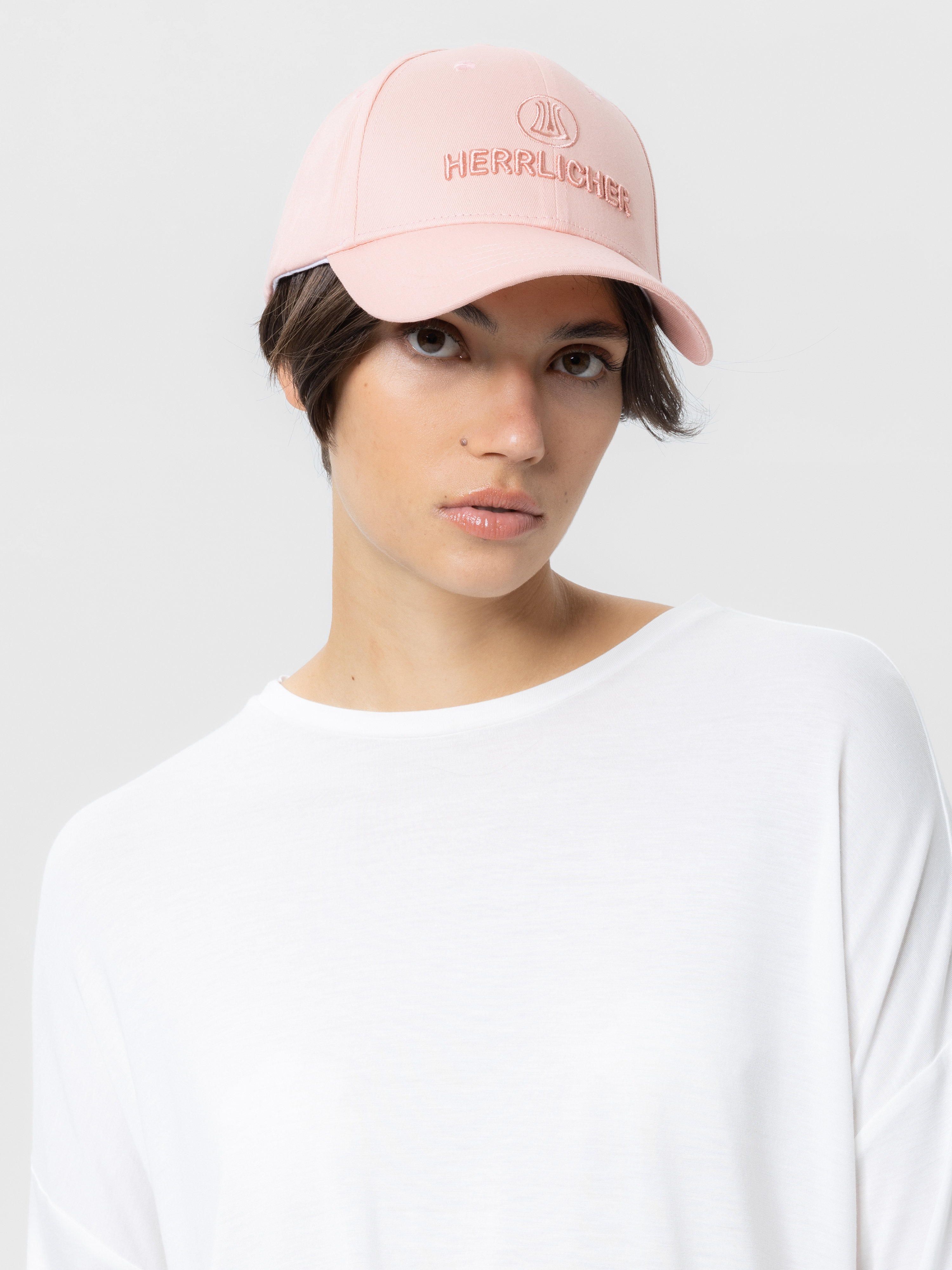 Eine Person, die eine hellrosa Herrlicher-Baseballmütze und ein lockeres weißes Oberteil trägt, blickt mit neutralem Gesichtsausdruck in die Kamera vor einem schlichten hellen Hintergrund.
