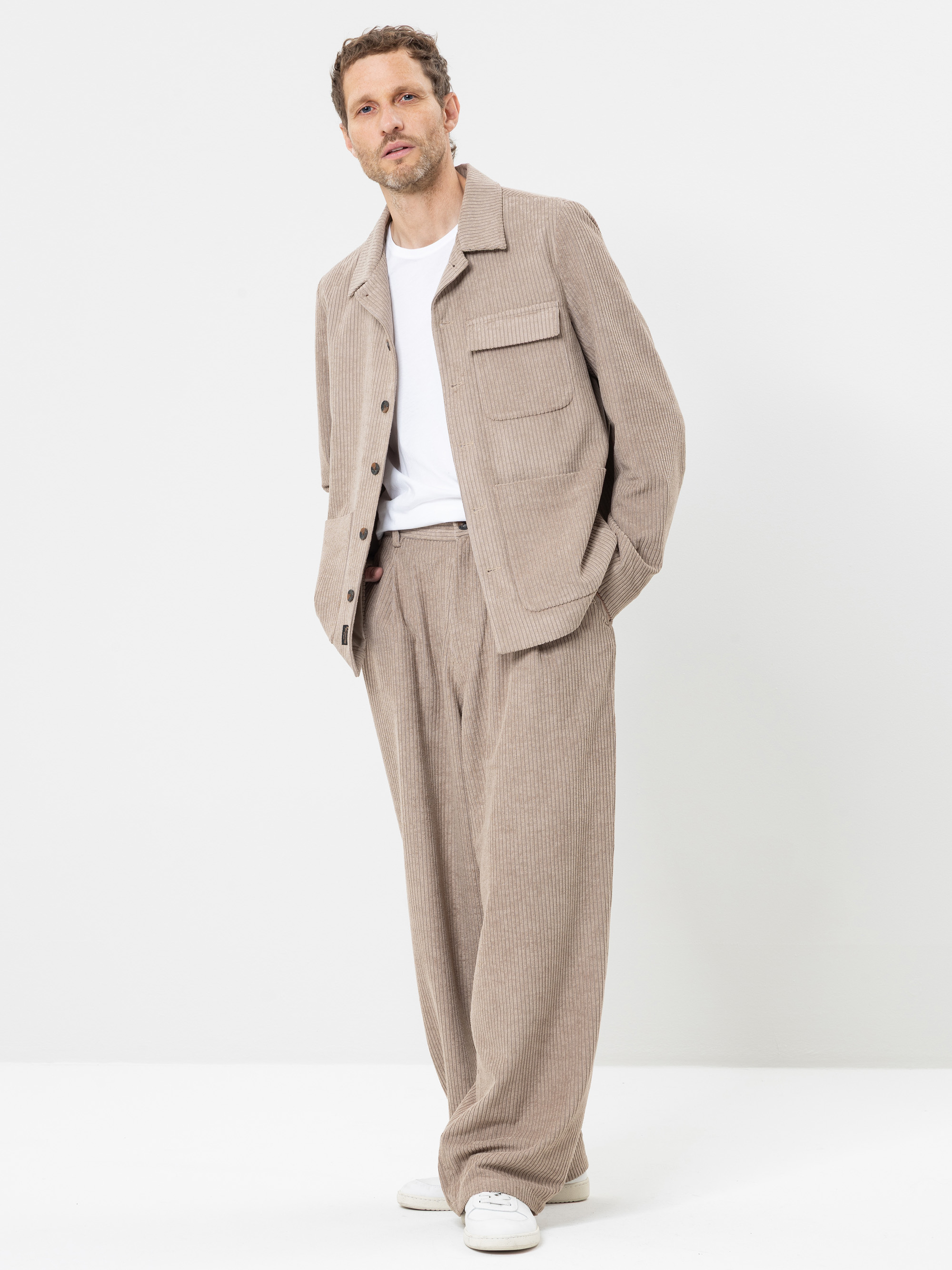 Ein Mann, der eine beige Cordjacke und eine dazu passende Hose mit weitem Bein über einem weißen T-Shirt und weißen Sneakers trägt, steht vor einem schlichten weißen Hintergrund, die Hände in den Taschen.