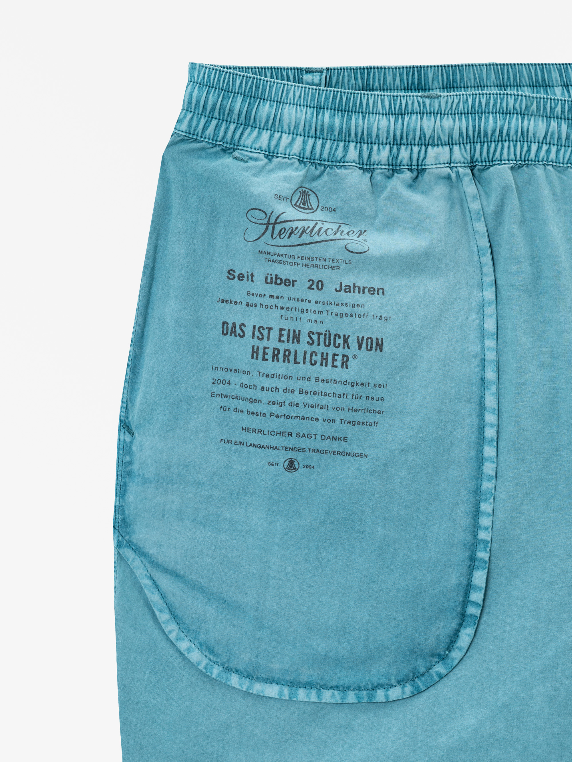 Nahaufnahme der Gesäßtasche einer hellblauen Hose mit deutschem Text und einem auf den Stoff über der Tasche gedruckten Logo, das die Marke Herrlicher und ihre Werte beschreibt.