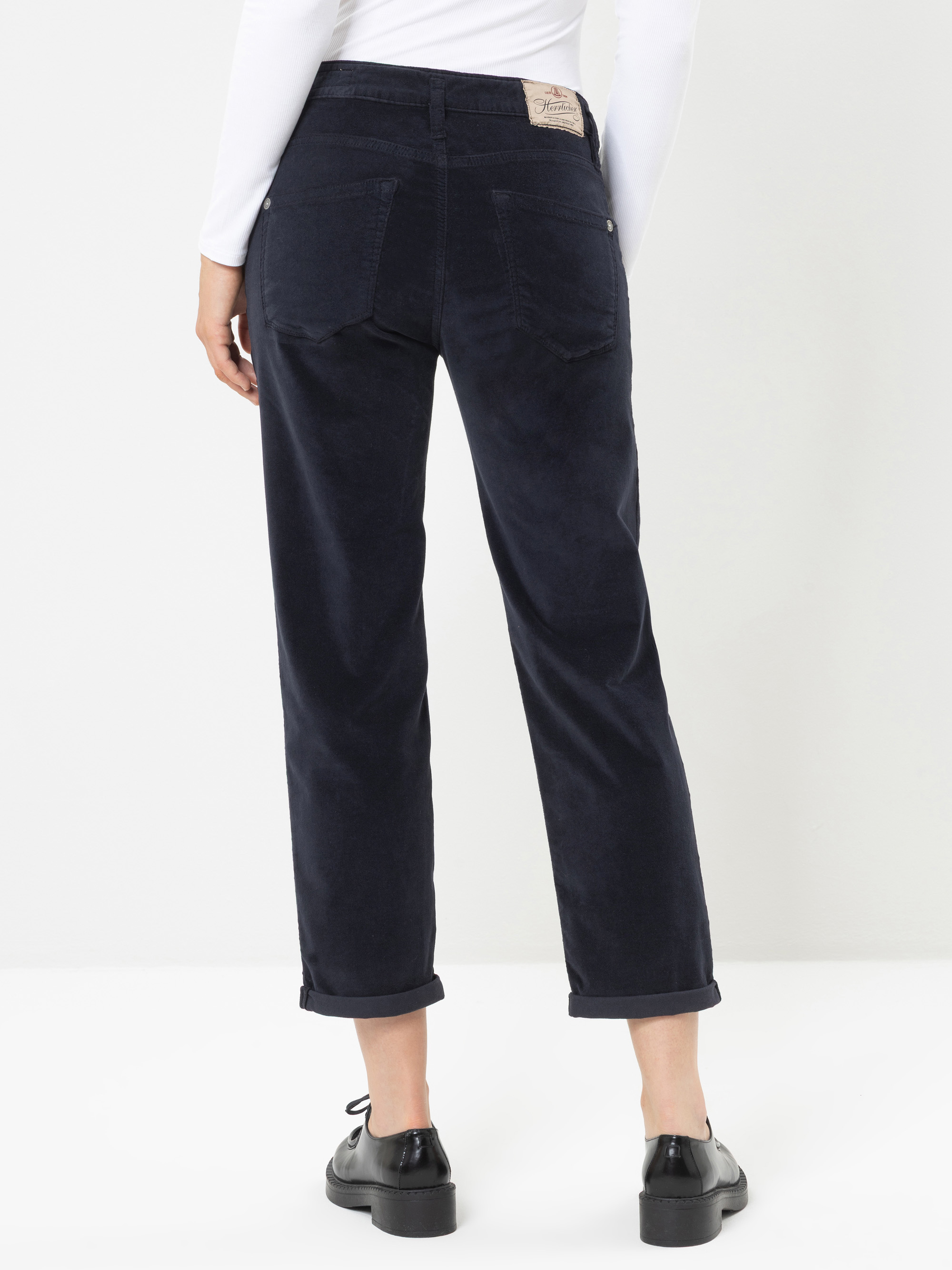Eine Person, die eine dunkelblaue Cordhose, ein weißes Langarmshirt und schwarze Schuhe trägt, steht mit dem Rücken zur Kamera, sodass die Gesäßtaschen und die hochgekrempelten Hosenbeine zu sehen sind.