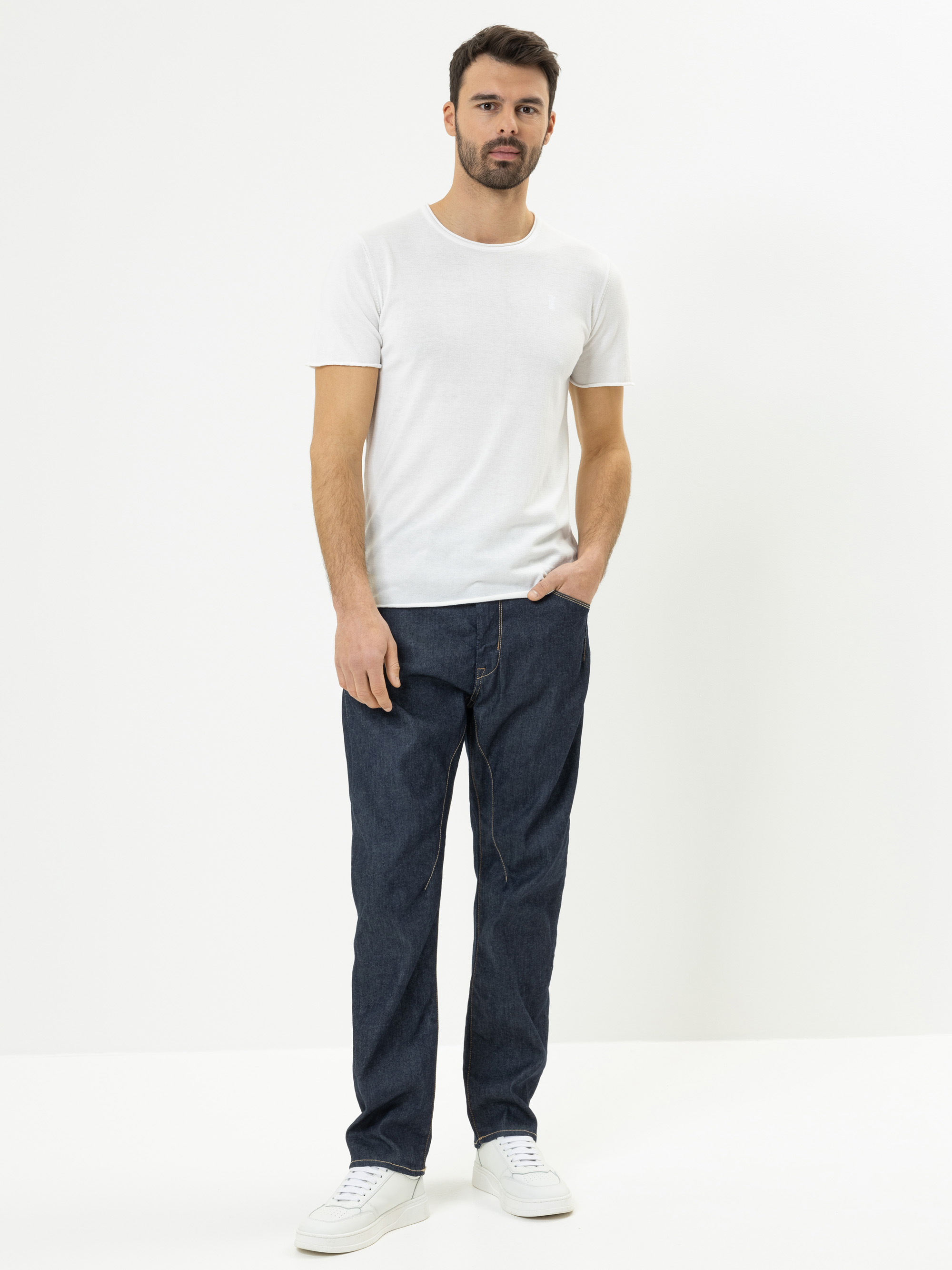 Ein Mann mit dunklem Haar und Bart steht vor einem schlichten weißen Hintergrund und trägt ein weißes T-Shirt, dunkelblaue Jeans und weiße Turnschuhe. Eine seiner Hände steckt in der Hosentasche.