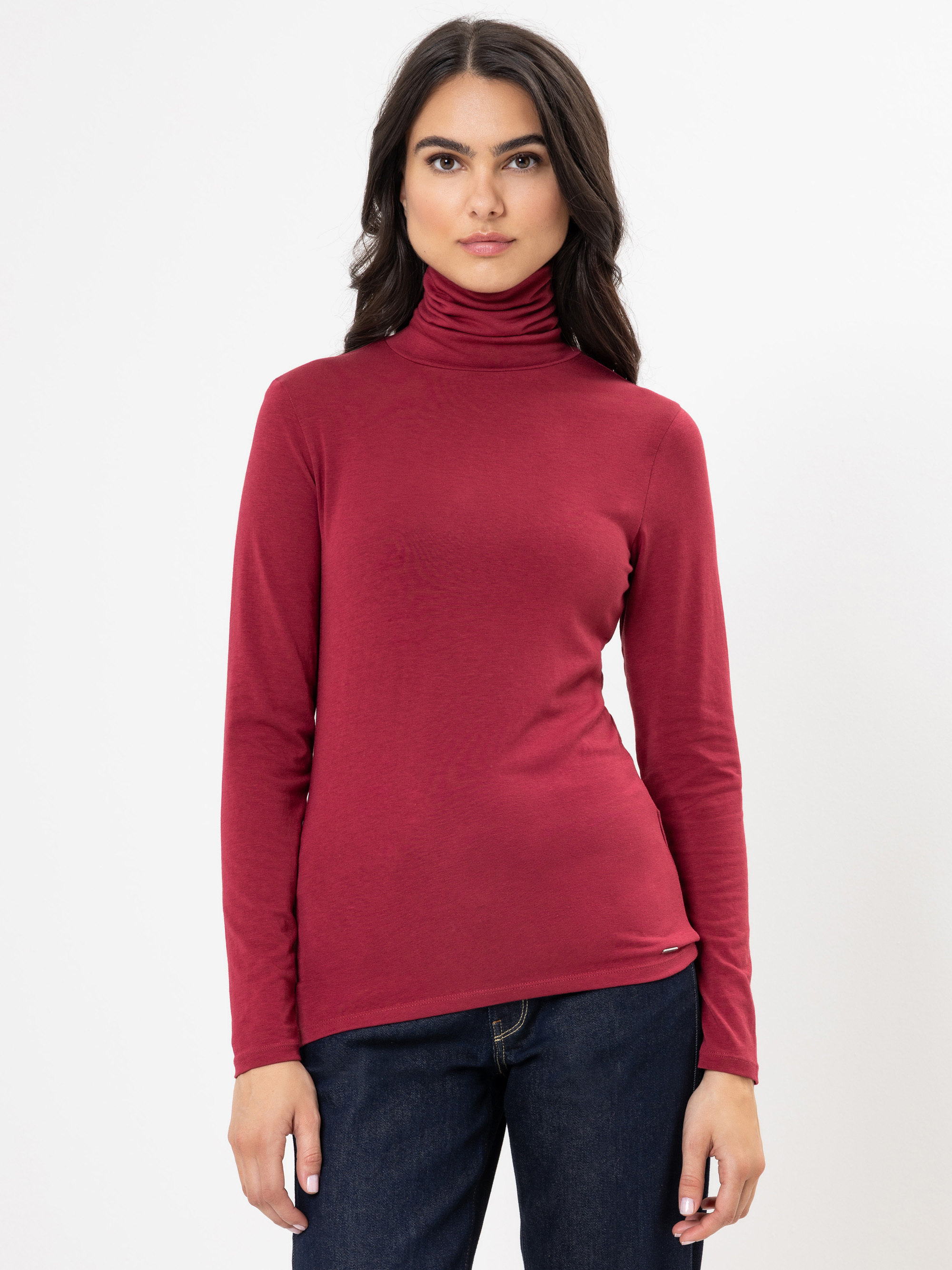 Eine Frau mit langen dunklen Haaren, die einen kastanienbraunen, langärmeligen Rollkragenpullover und dunkle Jeans trägt, steht vor einem schlichten weißen Hintergrund und blickt mit neutraler Miene nach vorn.