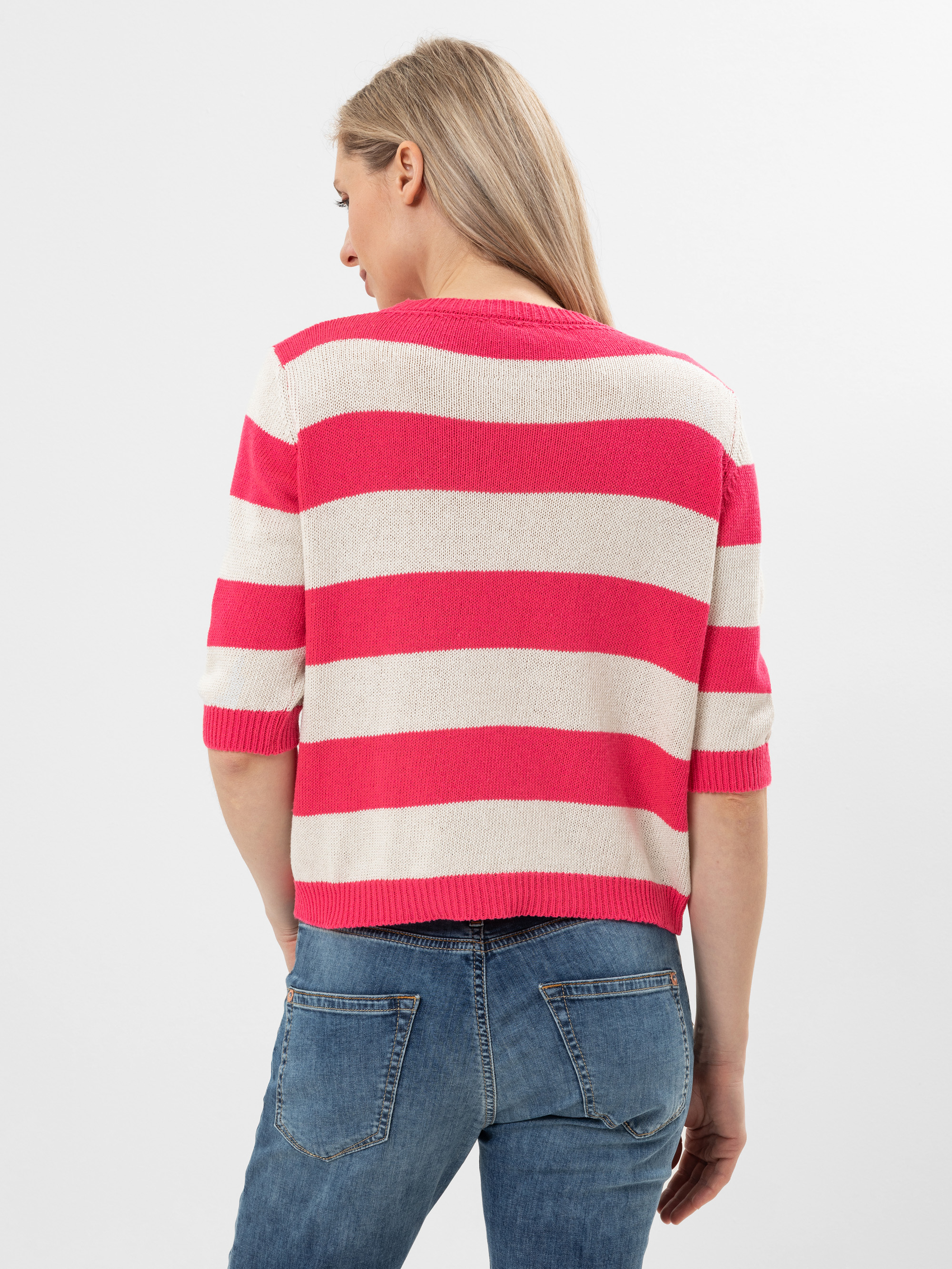 Eine Frau mit langen blonden Haaren ist von hinten zu sehen. Sie trägt einen rosa-weiß gestreiften Pullover mit ellenbogenlangen Ärmeln und blaue Jeans vor einem schlichten weißen Hintergrund.
