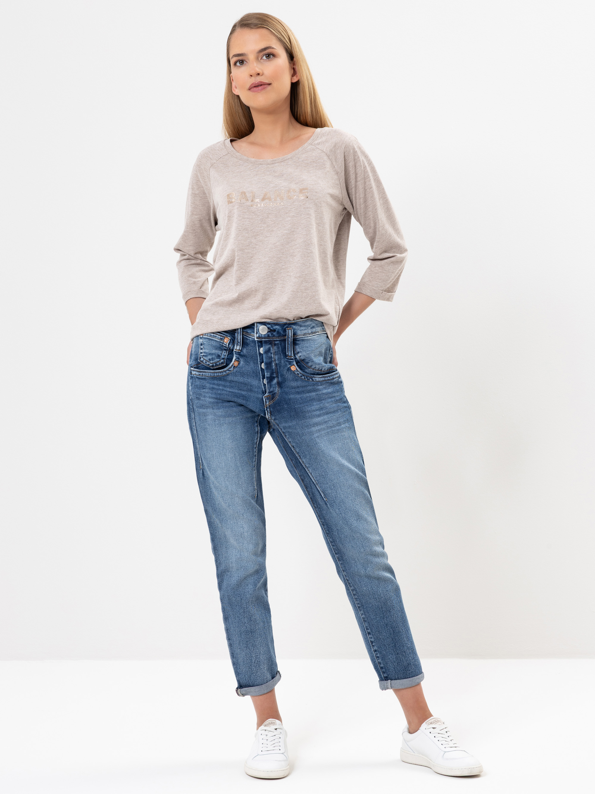 Eine Frau steht vor einem weißen Hintergrund und trägt ein hellbeiges langärmeliges Oberteil, blaue Jeans mit Bündchen und weiße Turnschuhe. Sie hat glattes, langes Haar und einen neutralen Gesichtsausdruck.