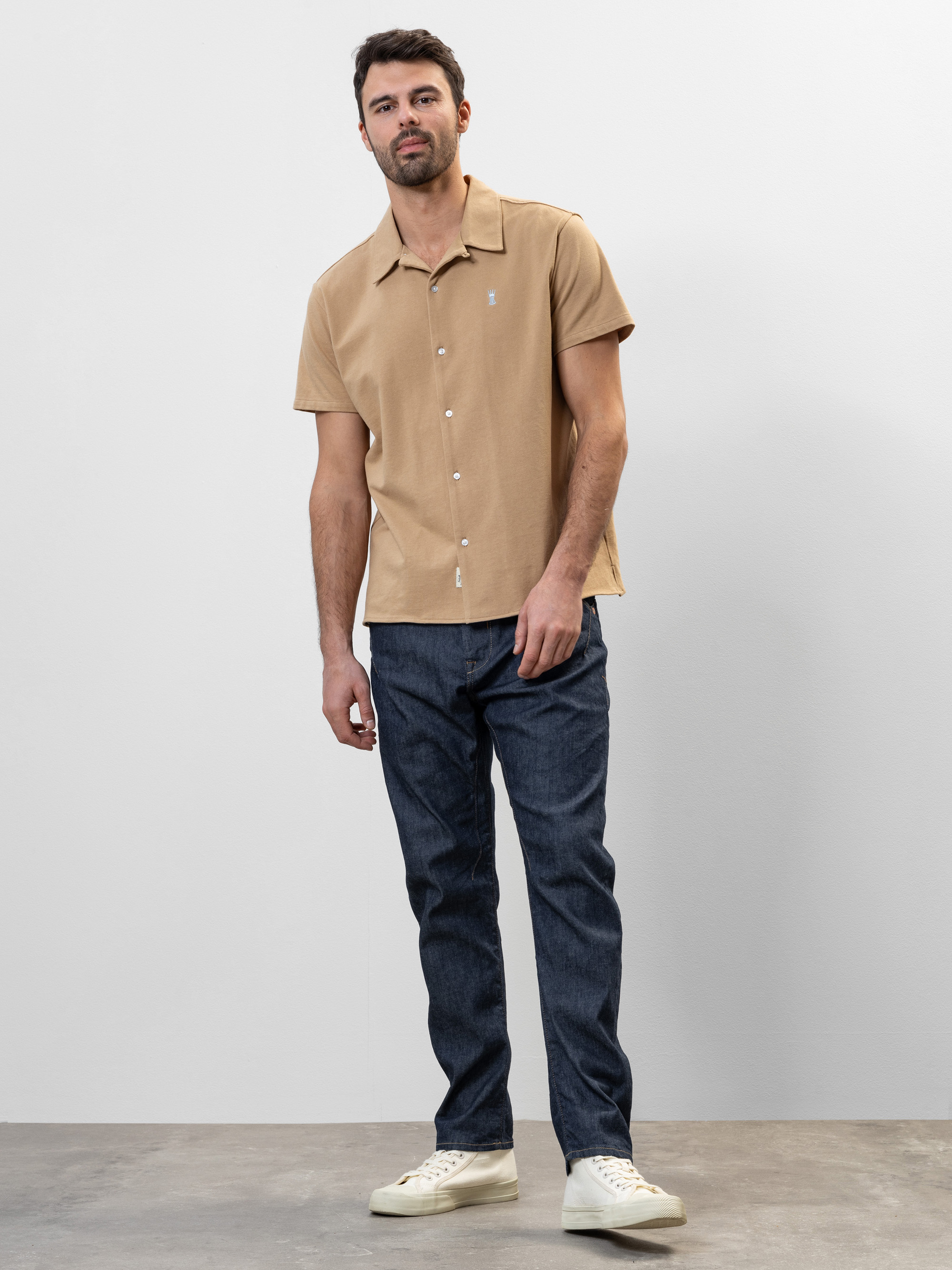 Ein Mann steht vor einem schlichten Hintergrund und trägt ein hellbraunes, kurzärmeliges Button-up-Hemd, dunkelblaue Jeans und weiße Turnschuhe.