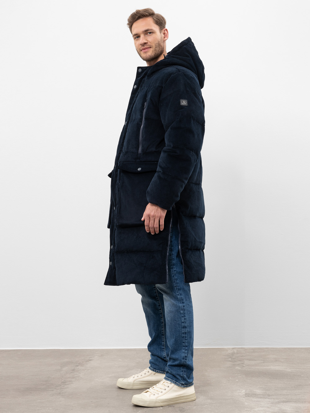 Ein Mann steht seitlich vor einem schlichten Hintergrund und trägt einen langen, dunklen Wintermantel mit Kapuze, blaue Jeans und weiße Turnschuhe.