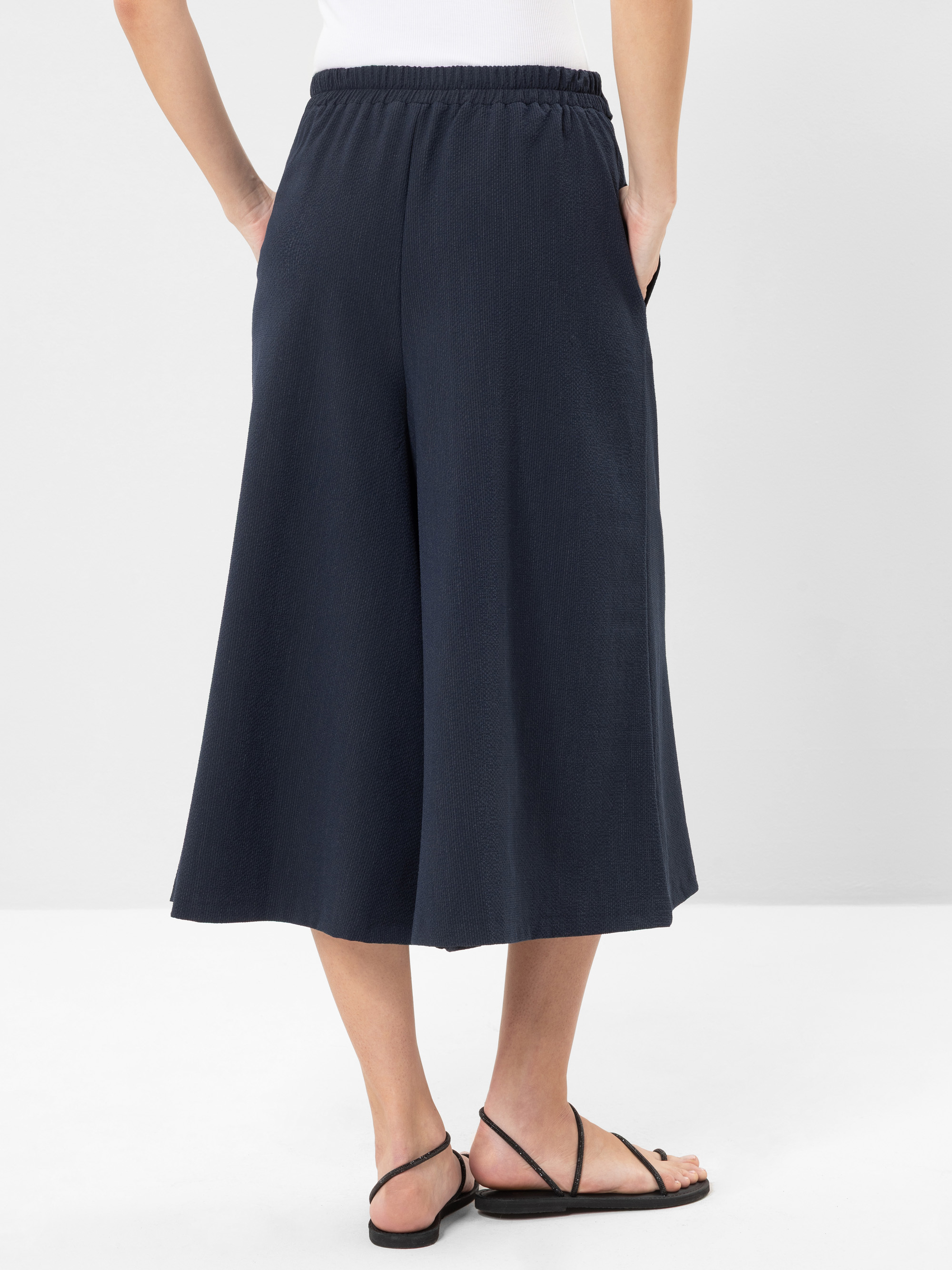 Person in lockerer, marineblauer Culotte-Hose, weißem Oberteil und schwarzen Sandalen, stehend mit den Händen in den Taschen und mit dem Rücken zur Kamera vor einem schlichten hellen Hintergrund.