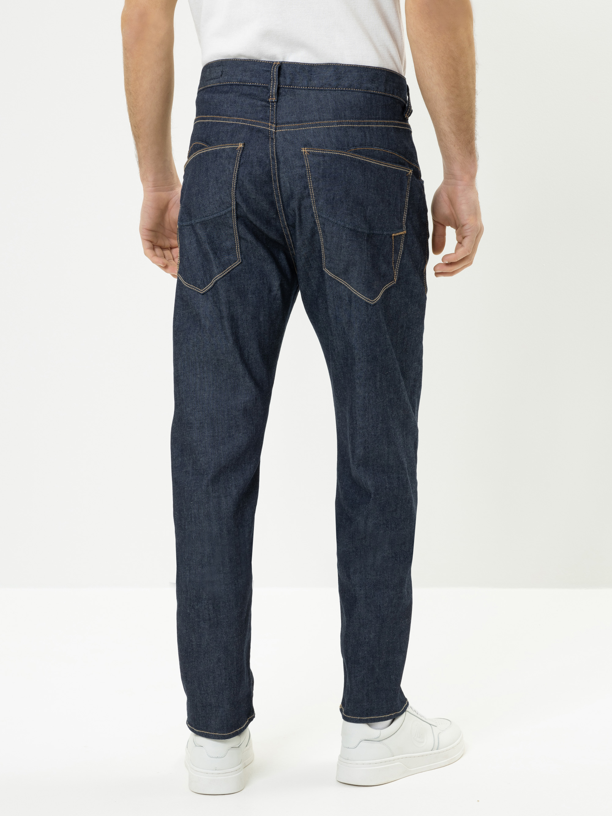 Eine Person, die ein weißes Hemd, eine dunkelblaue Jeans mit sichtbaren Nähten und weiße Turnschuhe trägt, steht mit dem Gesicht zur Kamera vor einem schlichten weißen Hintergrund.