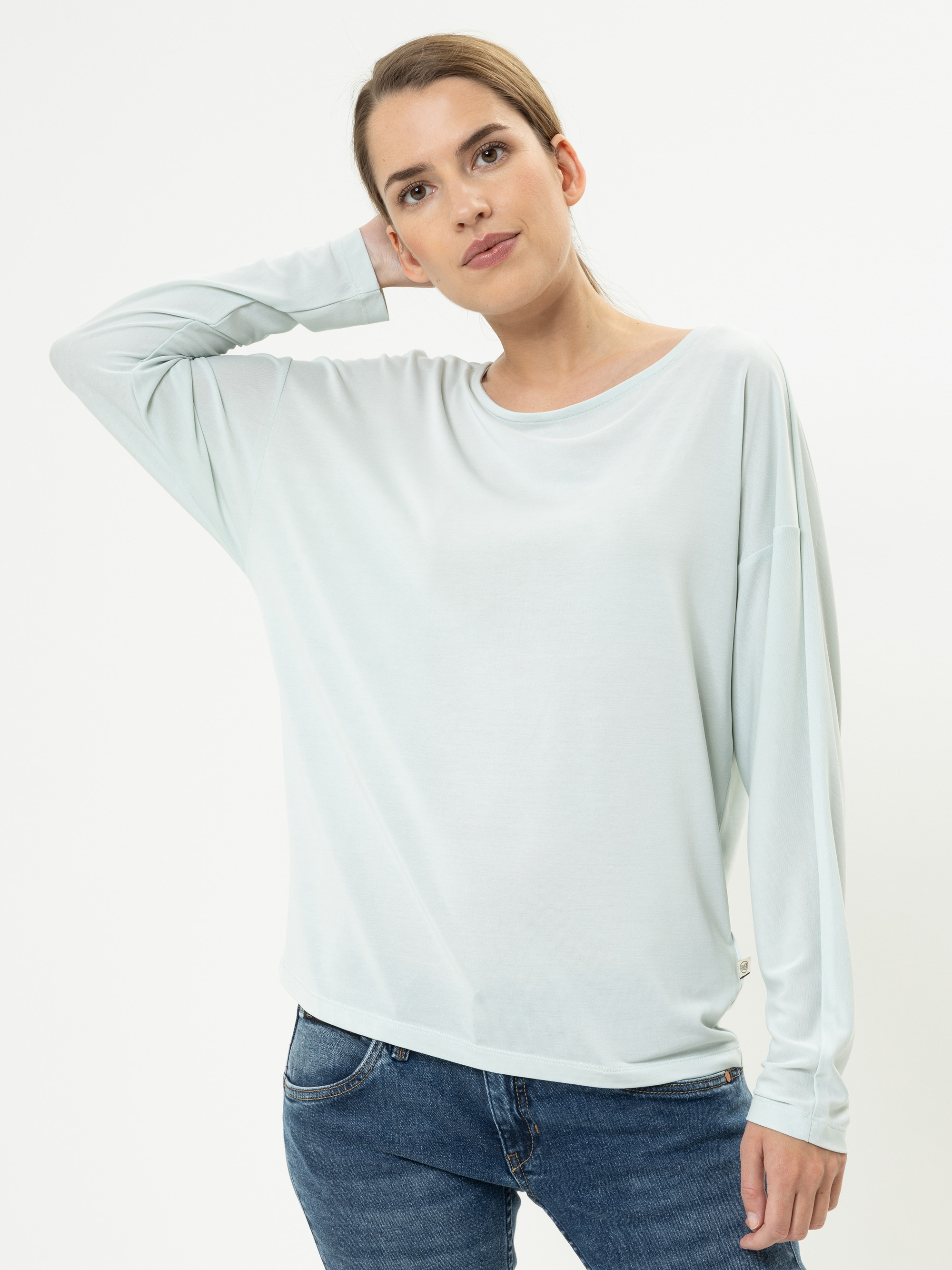Eine Frau mit hellbraunem, zurückgebundenem Haar trägt ein lockeres, helles, langärmeliges Hemd und blaue Jeans. Sie steht vor einem weißen Hintergrund und hat eine Hand hinter dem Kopf verschränkt.