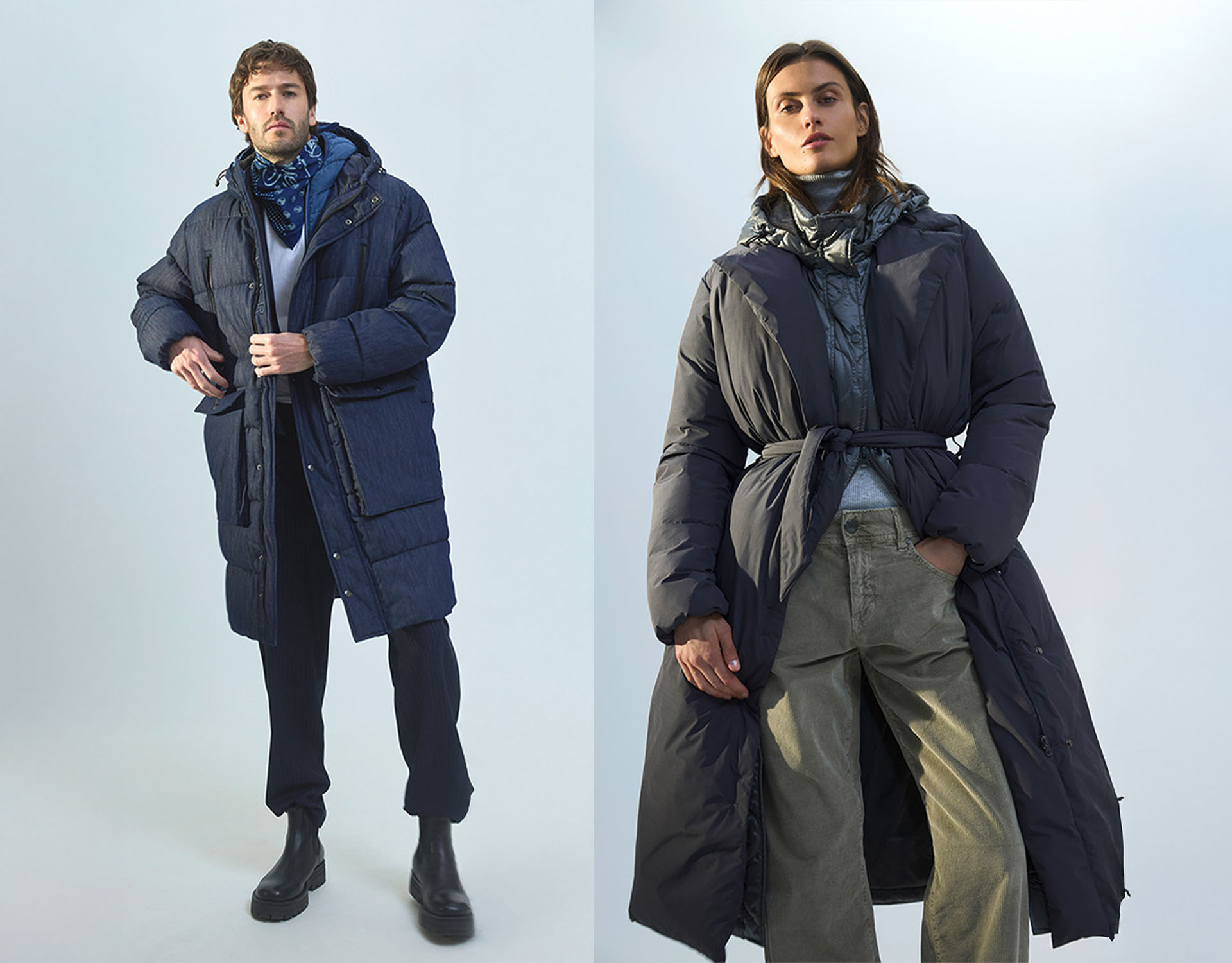 Ein Mann und eine Frau stehen nebeneinander und tragen jeweils einen langen, dunkelblauen Puffermantel mit Kapuze. Der Mann kombiniert seinen Mantel mit einer schwarzen Hose und Stiefeln, während die Frau eine helle Cargohose trägt und ihren Mantel in d
