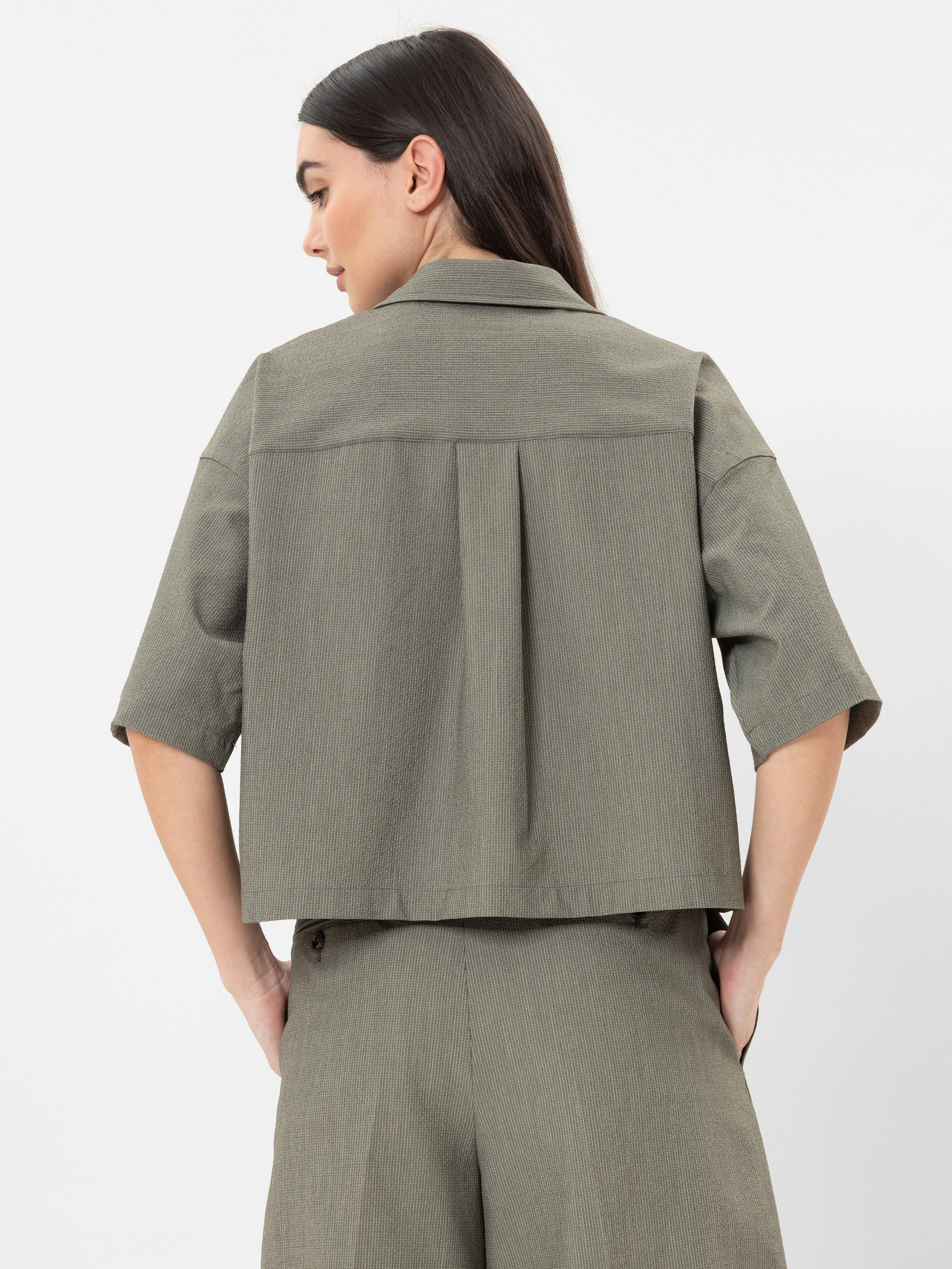 Eine Frau mit langen dunklen Haaren blickt von sich weg und trägt eine olivgrüne kurzärmelige Jacke und eine passende Hose. Die Jacke hat eine Rückenfalte und eine lockere, kastenförmige Passform. Sie steht vor einem schlichten weißen Hintergrund.