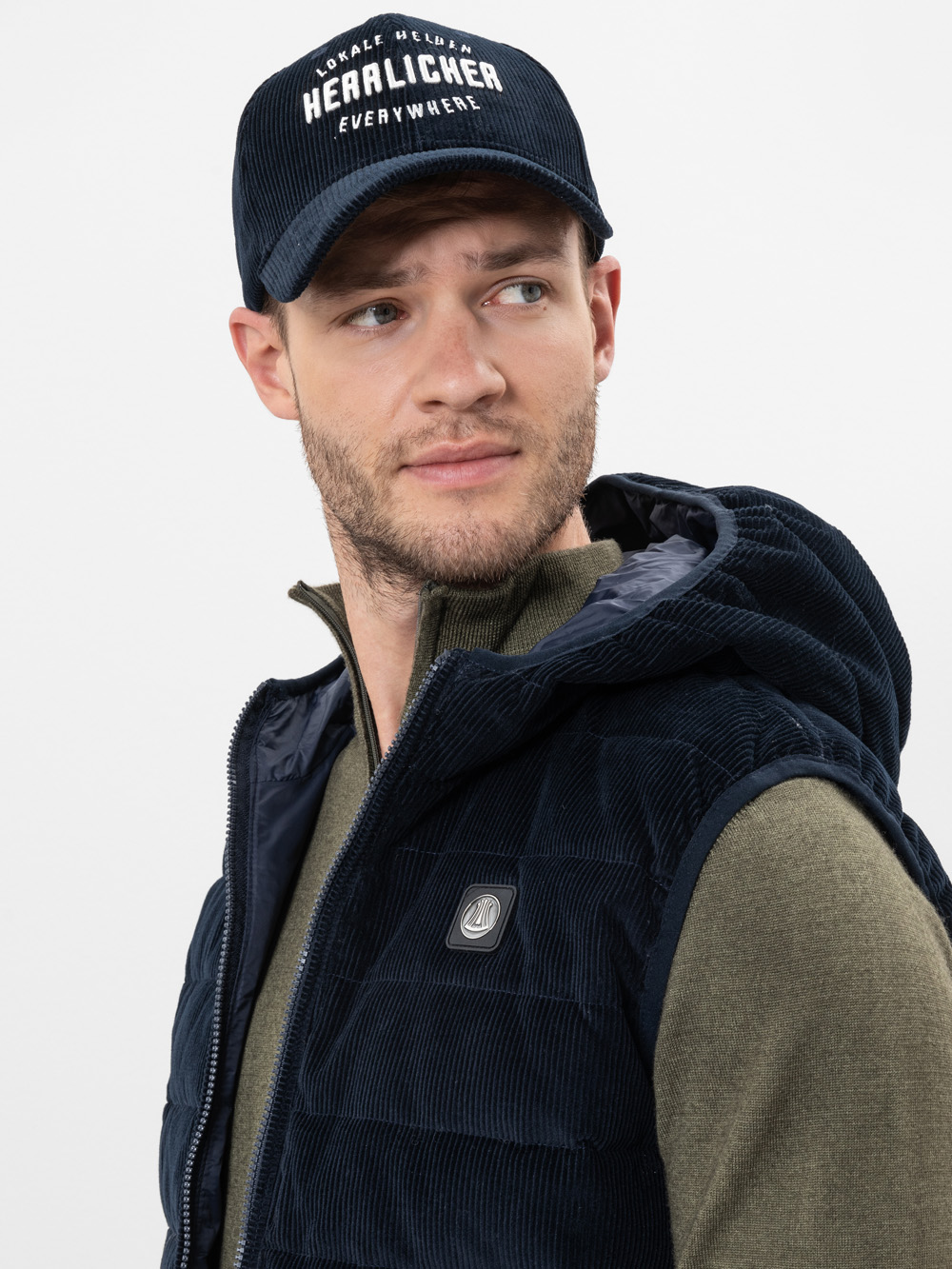 Ein Mann, der eine dunkelblaue Mütze mit weißer Aufschrift, eine passende blaue Steppweste und ein olivgrünes Hemd trägt, blickt vor einem schlichten weißen Hintergrund leicht nach links.