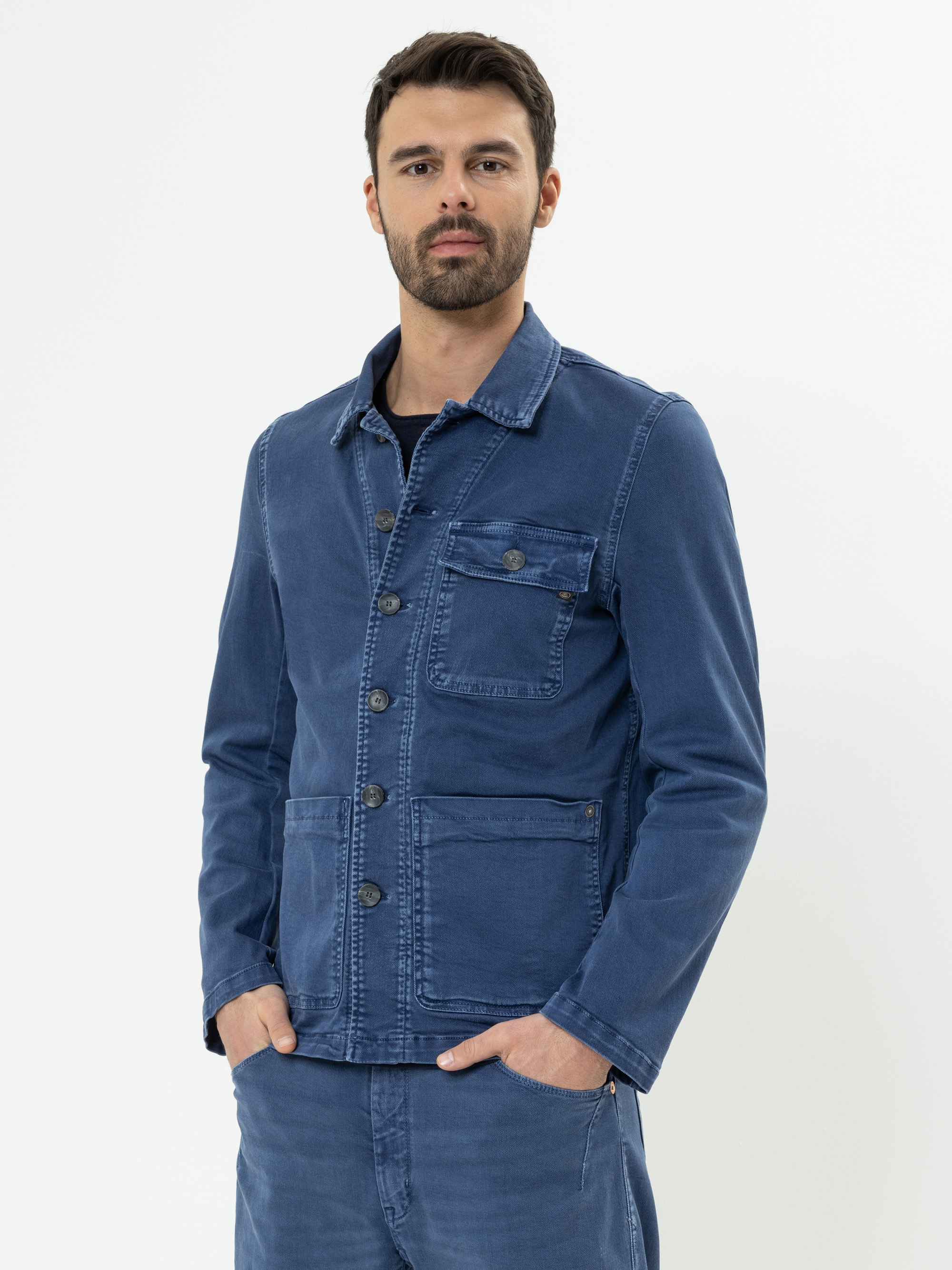 Ein Mann mit kurzen dunklen Haaren und einem Bart trägt eine blaue Jacke mit großen Vordertaschen und eine dazu passende blaue Hose und steht vor einem schlichten weißen Hintergrund.