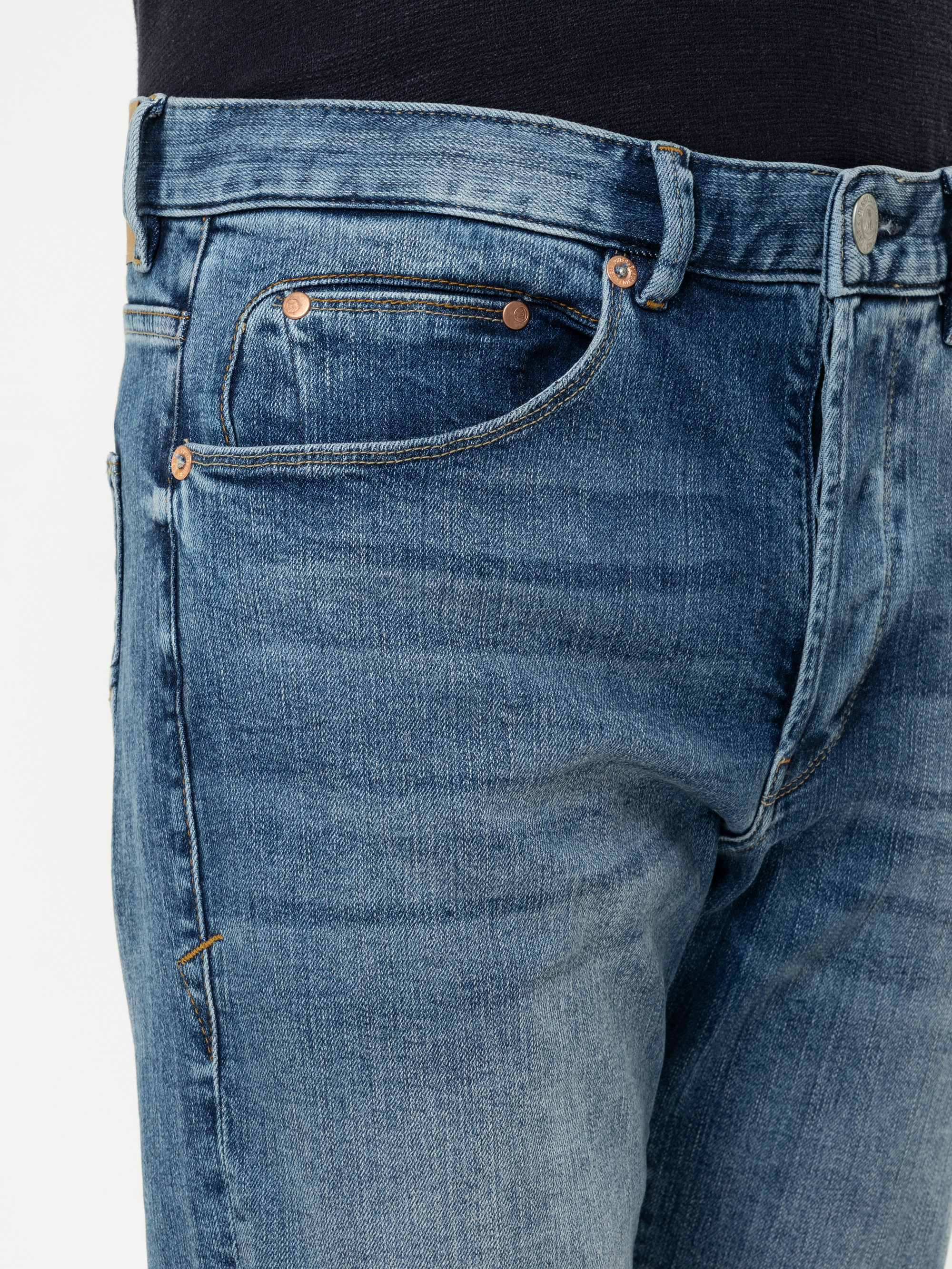 Nahaufnahme des oberen Teils einer blauen Denim-Jeans, die die Vordertasche, die Münztasche, den Bund und den Knopfverschluss zeigt. Die Jeans hat eine verblasste, abgenutzte Textur und kupferfarbene Nieten. Oben ist ein schwarzes Hemd teilweise sichtbar
