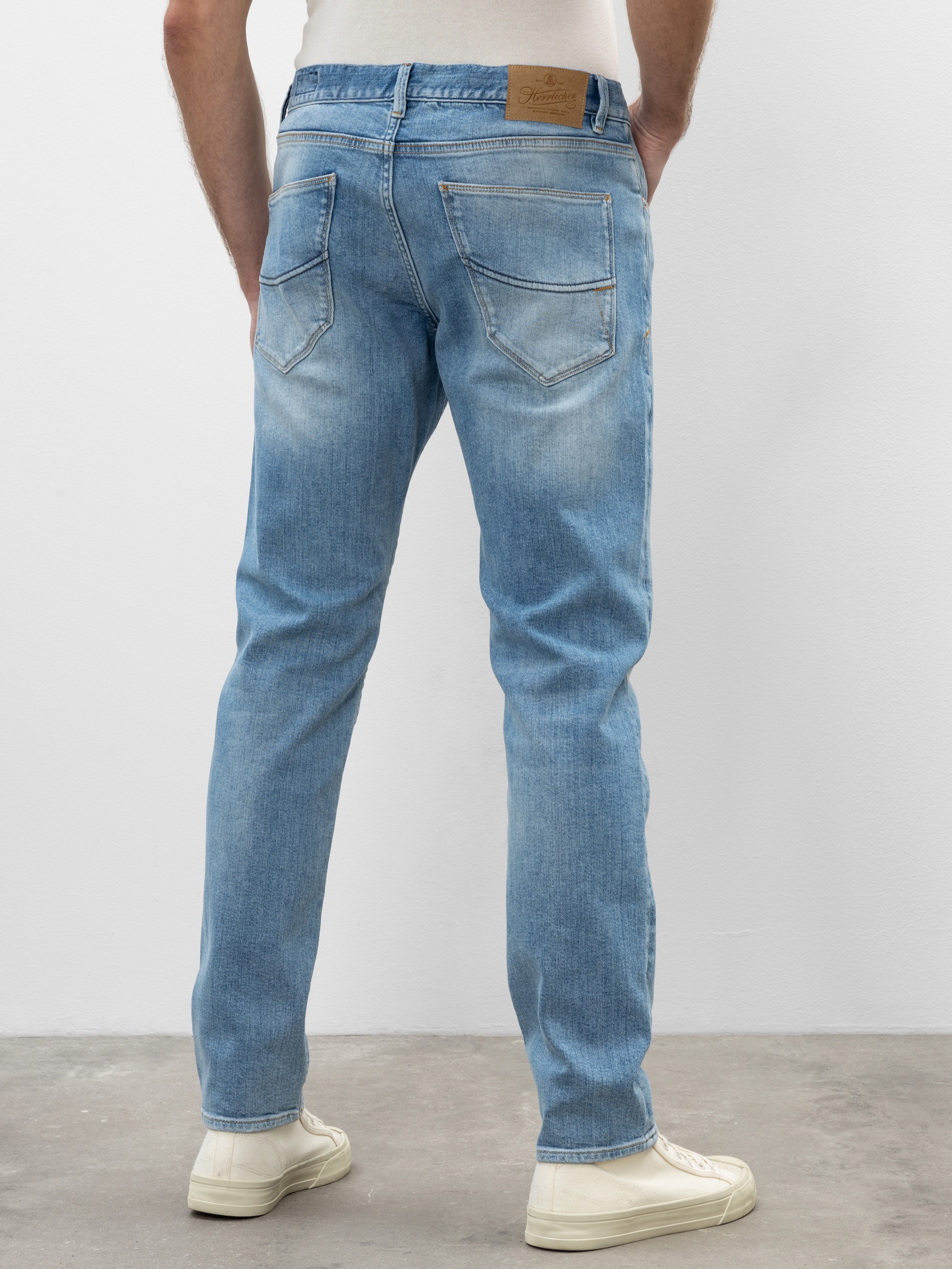 Eine Person in hellblauen Jeans und weißen Turnschuhen steht auf einem grauen Boden und blickt von der Kamera weg. Die Jeans hat verblasste Details und zwei Gesäßtaschen.