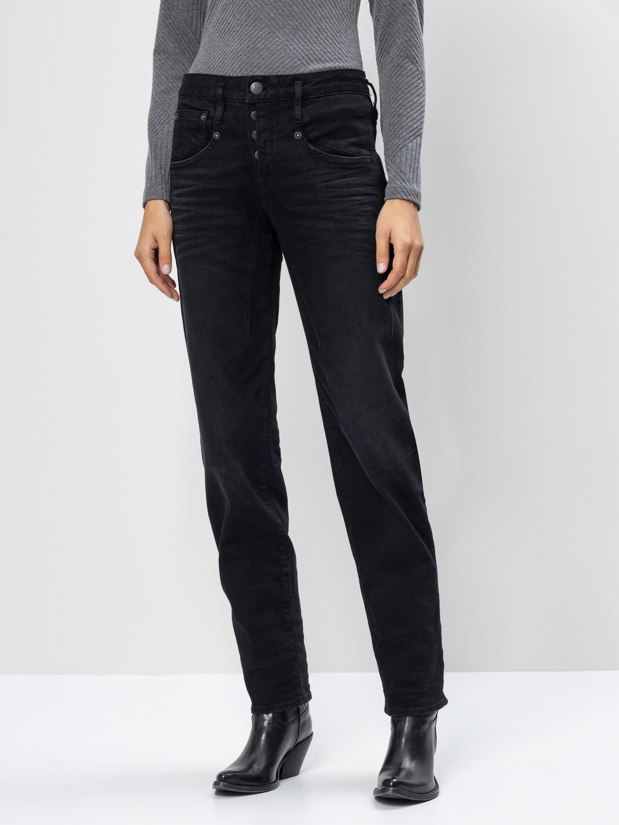 Eine Person, die ein langärmeliges, graues, geripptes Oberteil, eine schwarze, hoch taillierte Jeans mit mehreren Knöpfen und schwarze, hochhackige Stiefel trägt, steht vor einem schlichten, hellen Hintergrund.