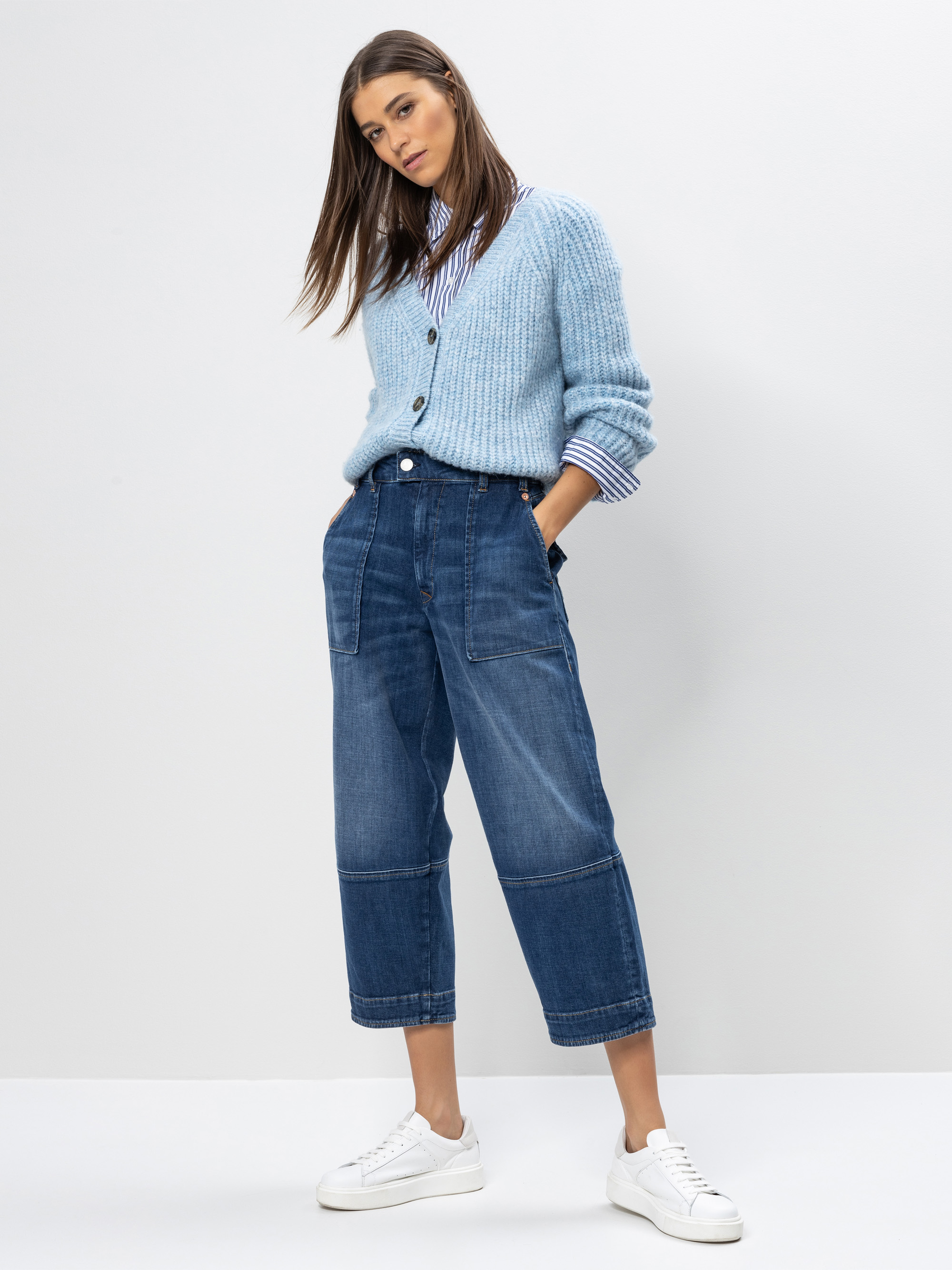 Eine Frau steht vor einem weißen Hintergrund und trägt eine hellblaue Strickjacke über einem gestreiften Hemd, weite Bluejeans und weiße Turnschuhe, die Hände in den Taschen und einen entspannten Gesichtsausdruck.