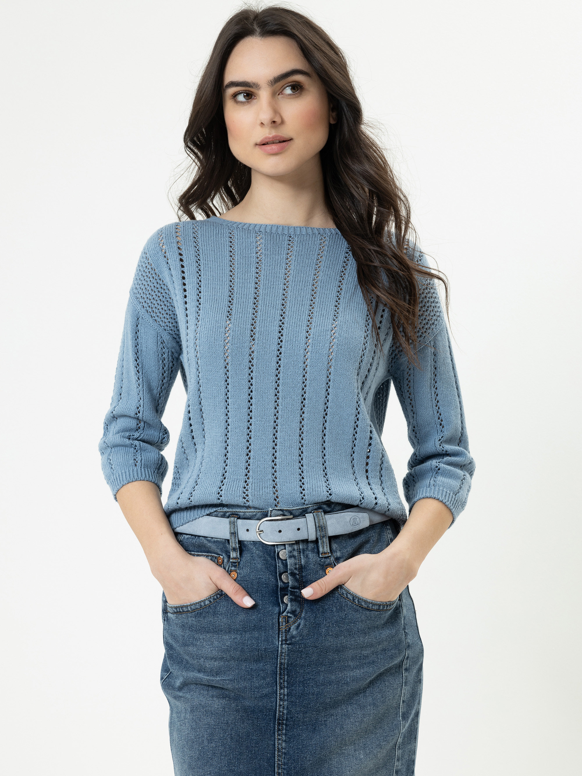 Eine Frau mit langen braunen Haaren trägt einen hellblauen Strickpullover und einen Jeansrock mit Knöpfen und steht mit den Händen in den Taschen vor einem schlichten weißen Hintergrund.