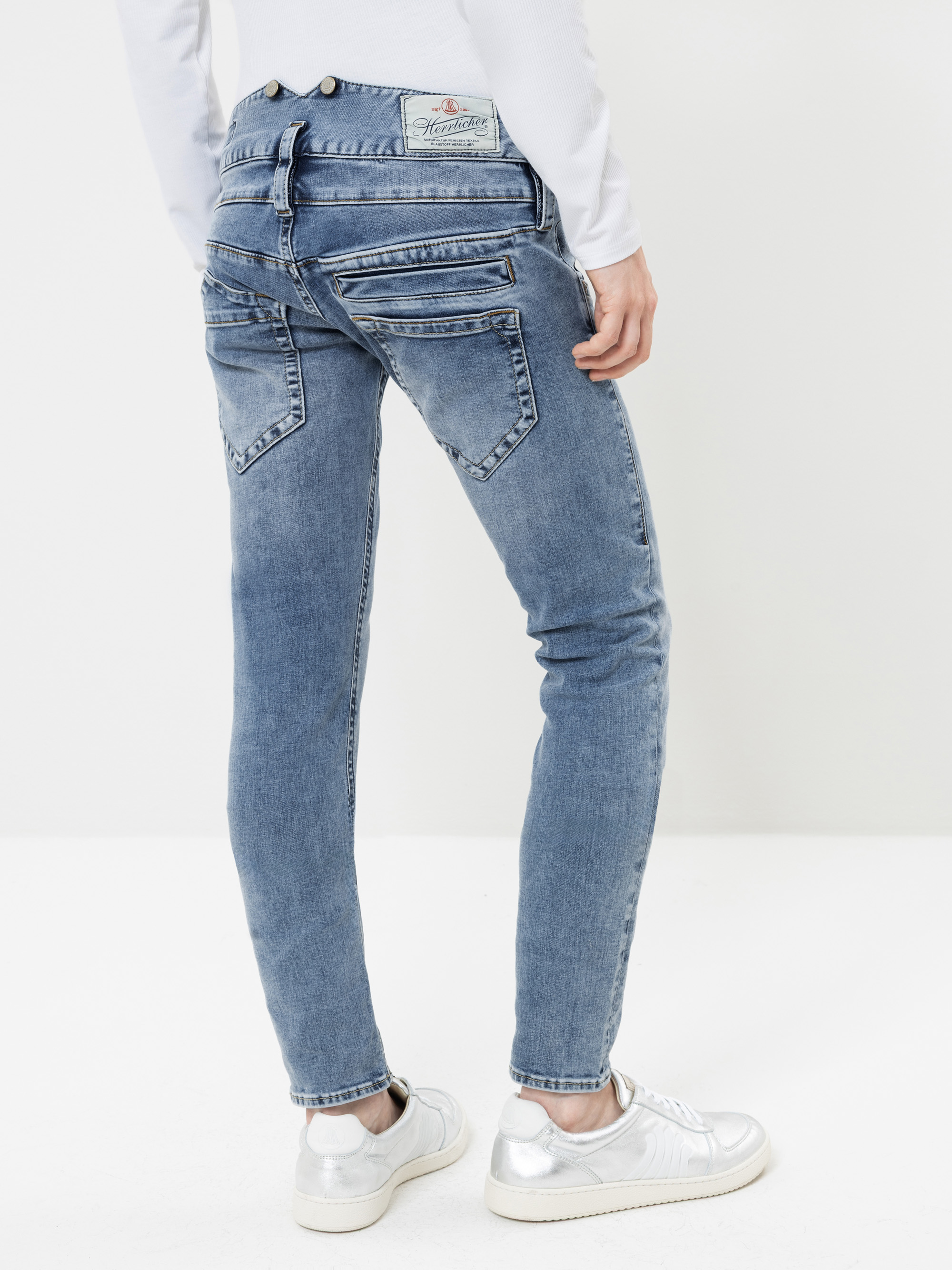 Eine Person, die eine blaue Skinny-Jeans mit mehreren Reißverschlüssen und Gesäßtaschen trägt, von hinten gesehen. Die Jeans wird mit einem weißen Oberteil und silbernen Sneakers kombiniert. Der Hintergrund ist schlicht weiß.
