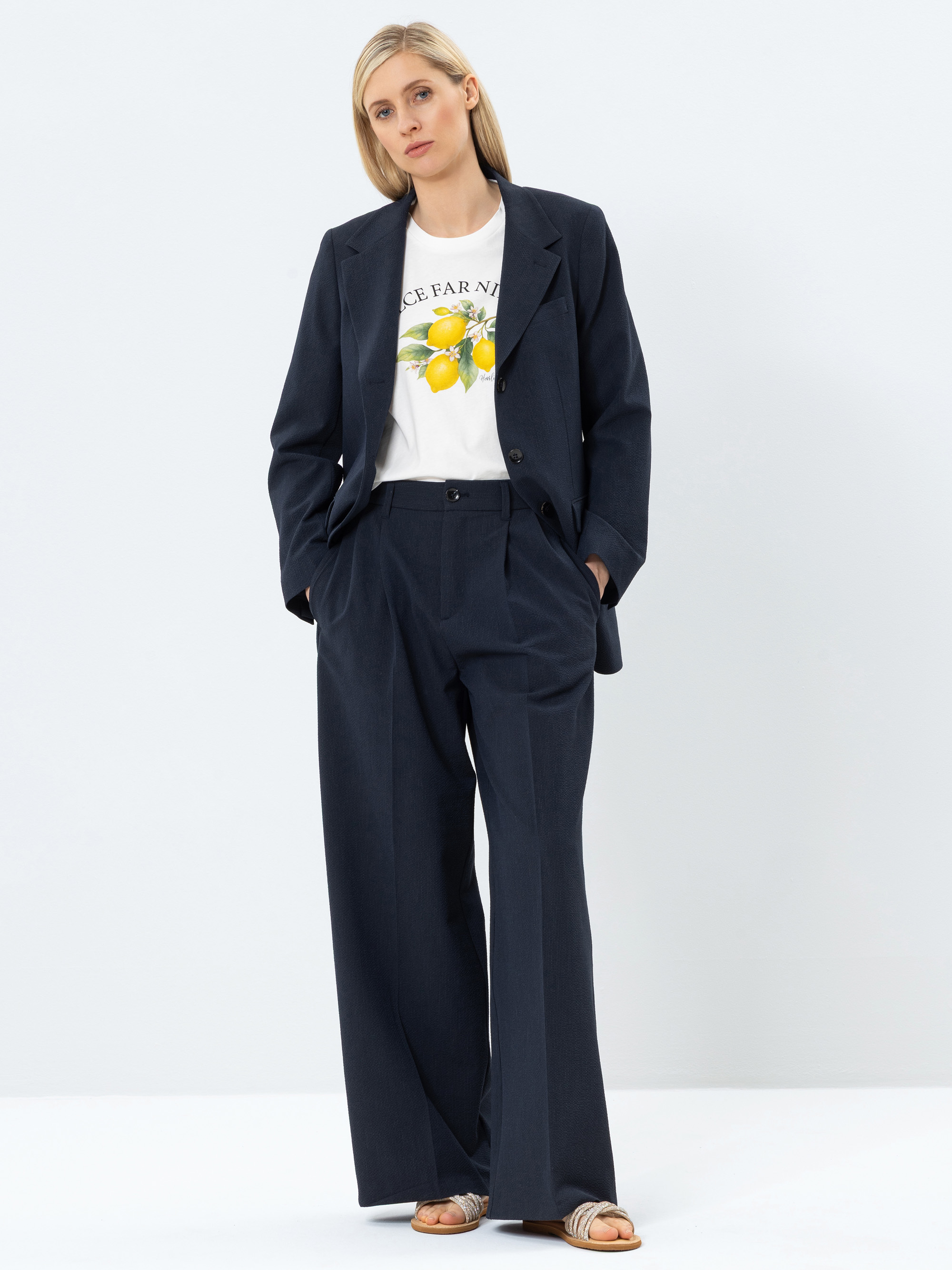 Eine Frau steht vor einem schlichten Hintergrund und trägt einen dunkeln marineblauen Anzug mit weiten Hosenbeinen, ein weißes T-Shirt mit einem Zitronenmotiv und flache Sandalen. Ihre Hände stecken in den Taschen und sie hat glattes, blondes Haar.