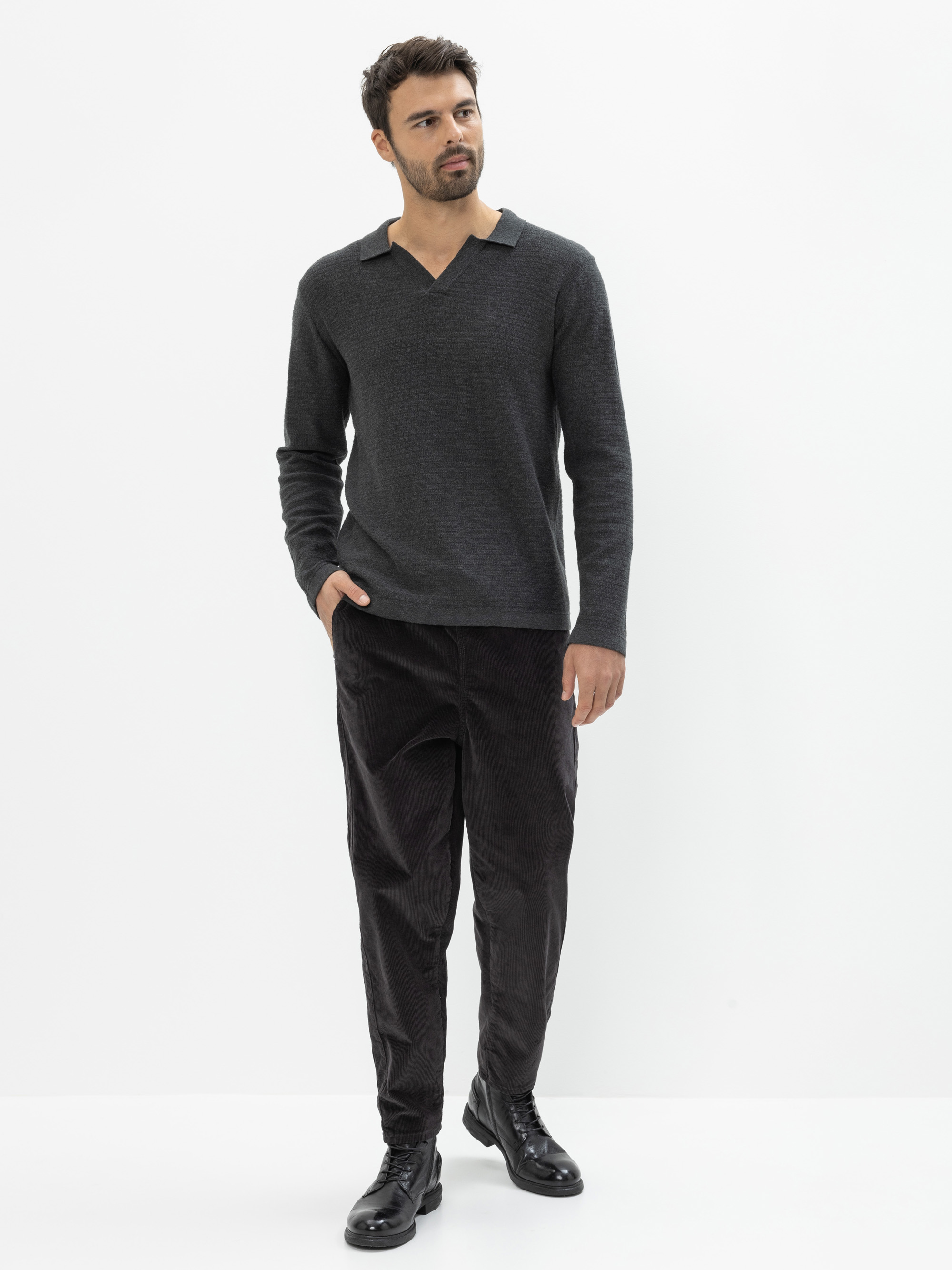 Ein Mann steht vor einem schlichten weißen Hintergrund, trägt ein dunkelgraues, langärmeliges Hemd mit V-Ausschnitt, eine schwarze Hose und schwarze Schuhe, hat eine Hand in der Tasche und schaut leicht zur Seite.