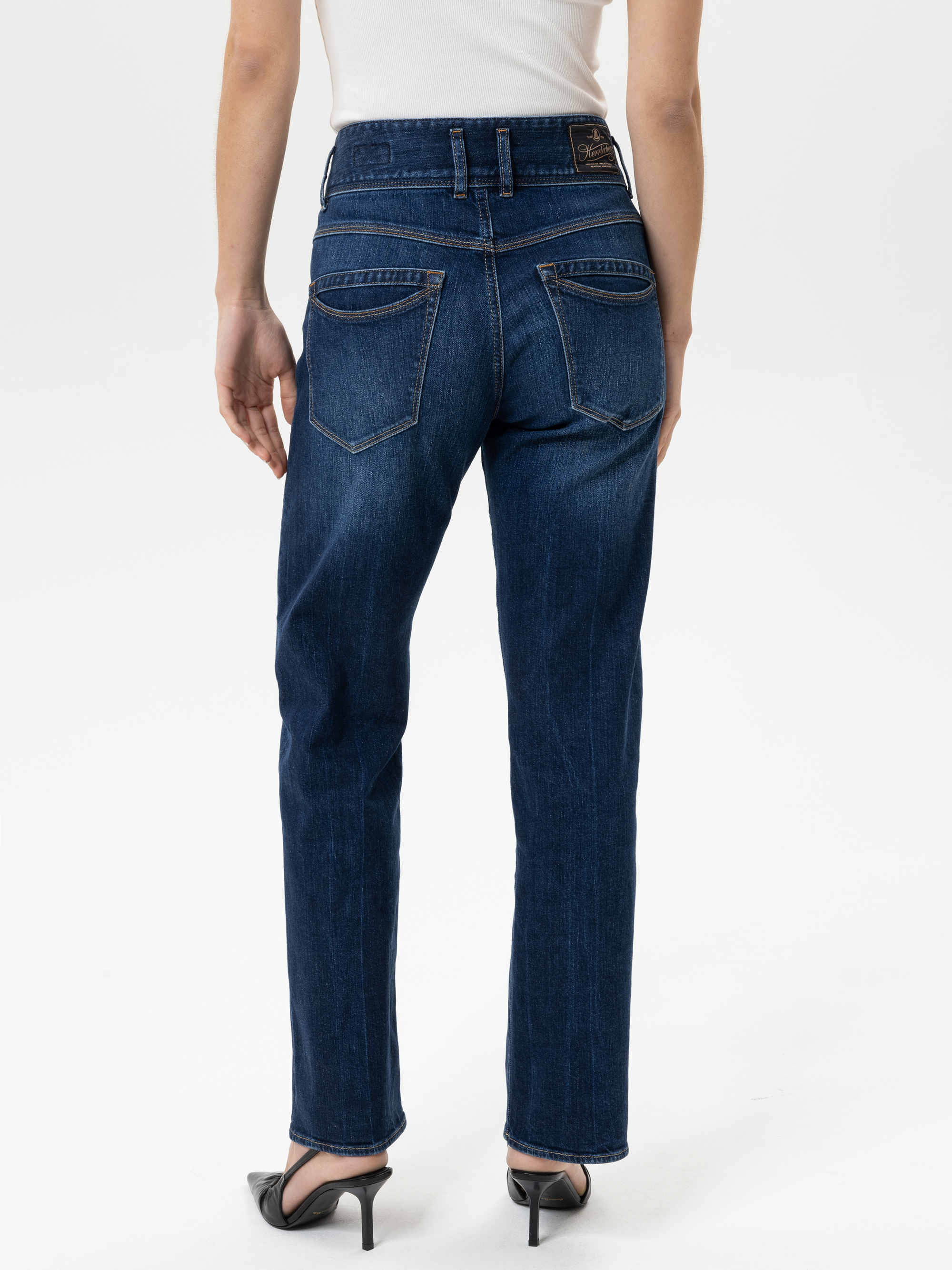 Eine Person, die ein weißes ärmelloses Oberteil, dunkelblaue Jeans mit geradem Bein und schwarze hochhackige Sandalen trägt, wird von hinten vor einem schlichten weißen Hintergrund gezeigt.