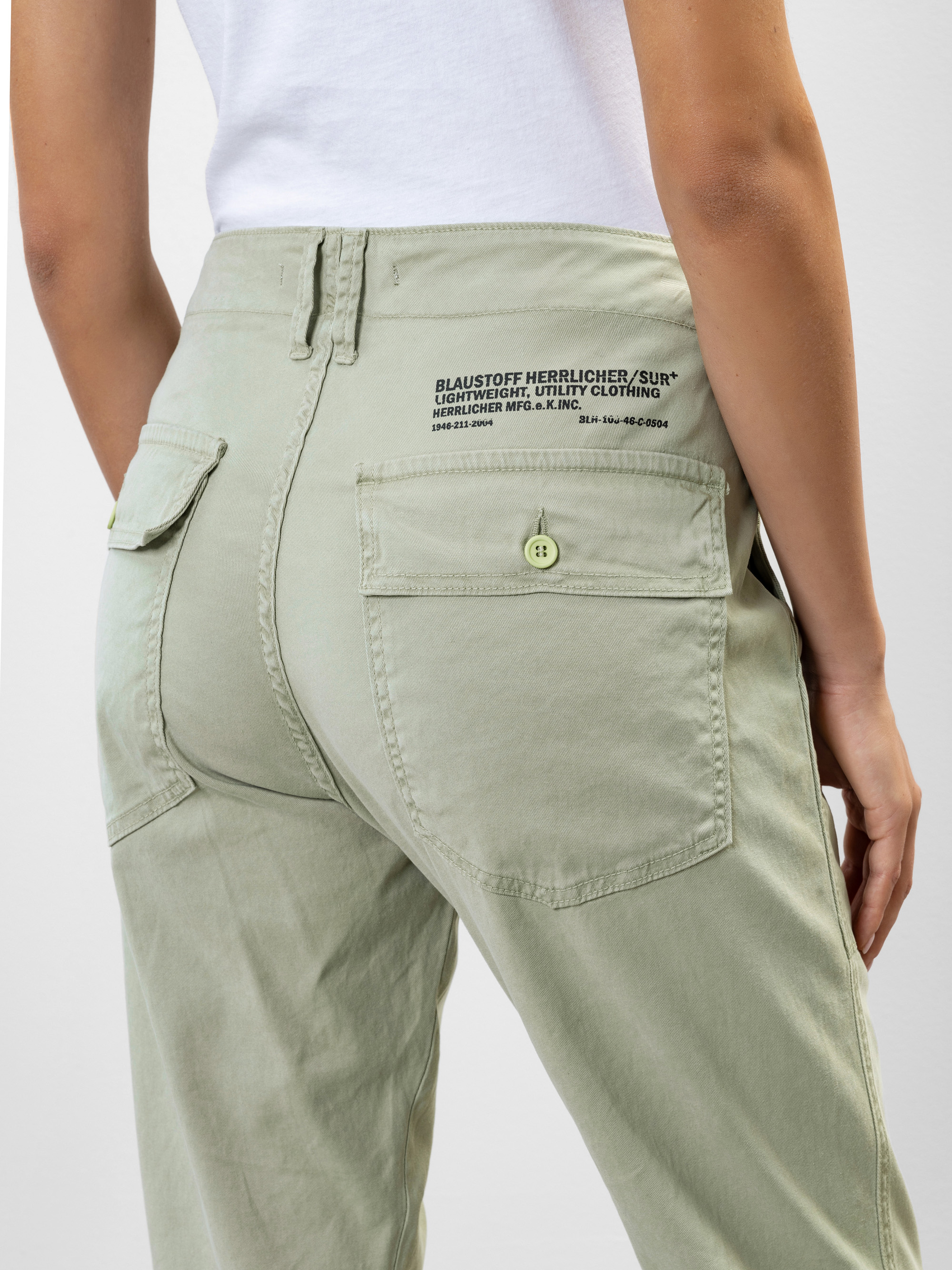 Eine Person, die eine hellgrüne Funktionshose mit einer großen Gesäßtasche mit schwarzem Aufdruck und einem Knopf trägt, dazu ein weißes T-Shirt, steht mit dem Rücken zur Kamera.