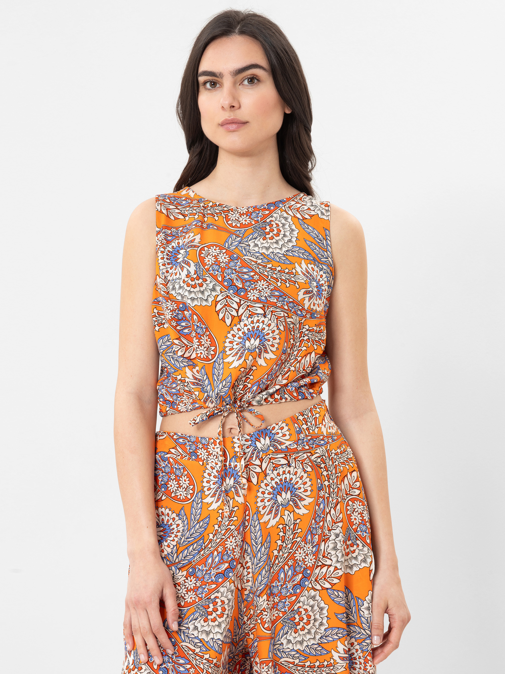 Eine Frau mit langen dunklen Haaren trägt ein ärmelloses orangefarbenes Oberteil und eine dazu passende Hose mit einem bunten Paisley- und Blumenmuster vor einem schlichten weißen Hintergrund.