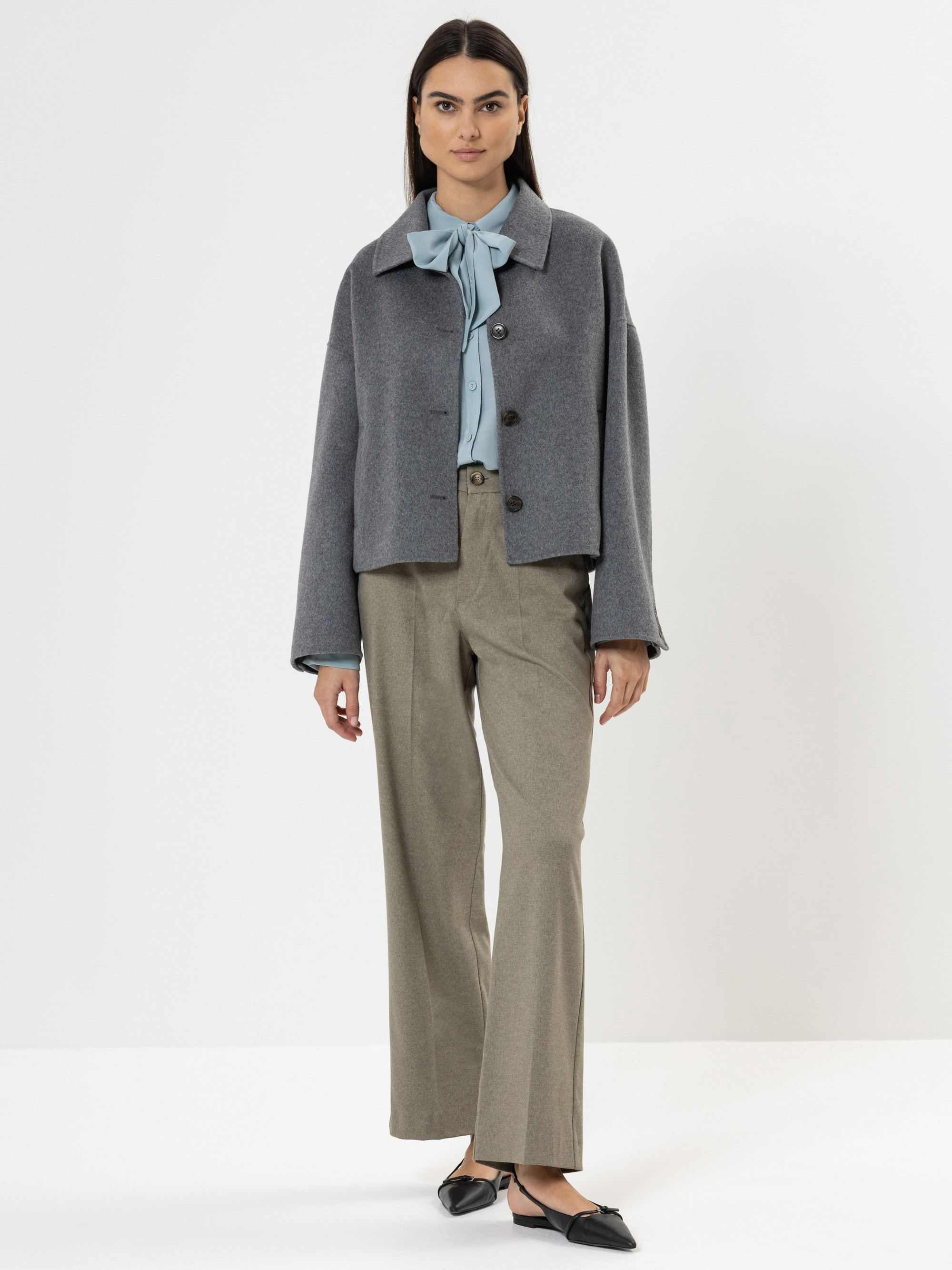 Eine Frau steht vor einem weißen Hintergrund und trägt eine graue Jacke über einer hellblauen Bluse mit Schleife, eine beigefarbene Hose mit weitem Bein und schwarze Schuhe mit spitzen Zehen. Sie hat glattes dunkles Haar und einen neutralen Gesichtsaus