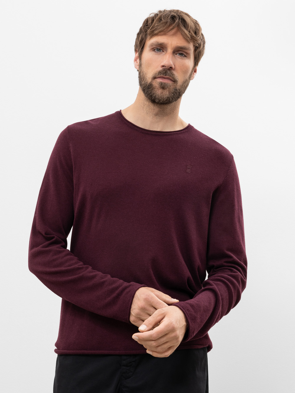 Ein Mann mit kurzen braunen Haaren und einem Bart trägt einen langärmeligen, weinroten Pullover und eine schwarze Hose. Er steht vor einem schlichten weißen Hintergrund und blickt leicht zur Seite, wobei er eine Hand in die Nähe seiner Taille stützt.