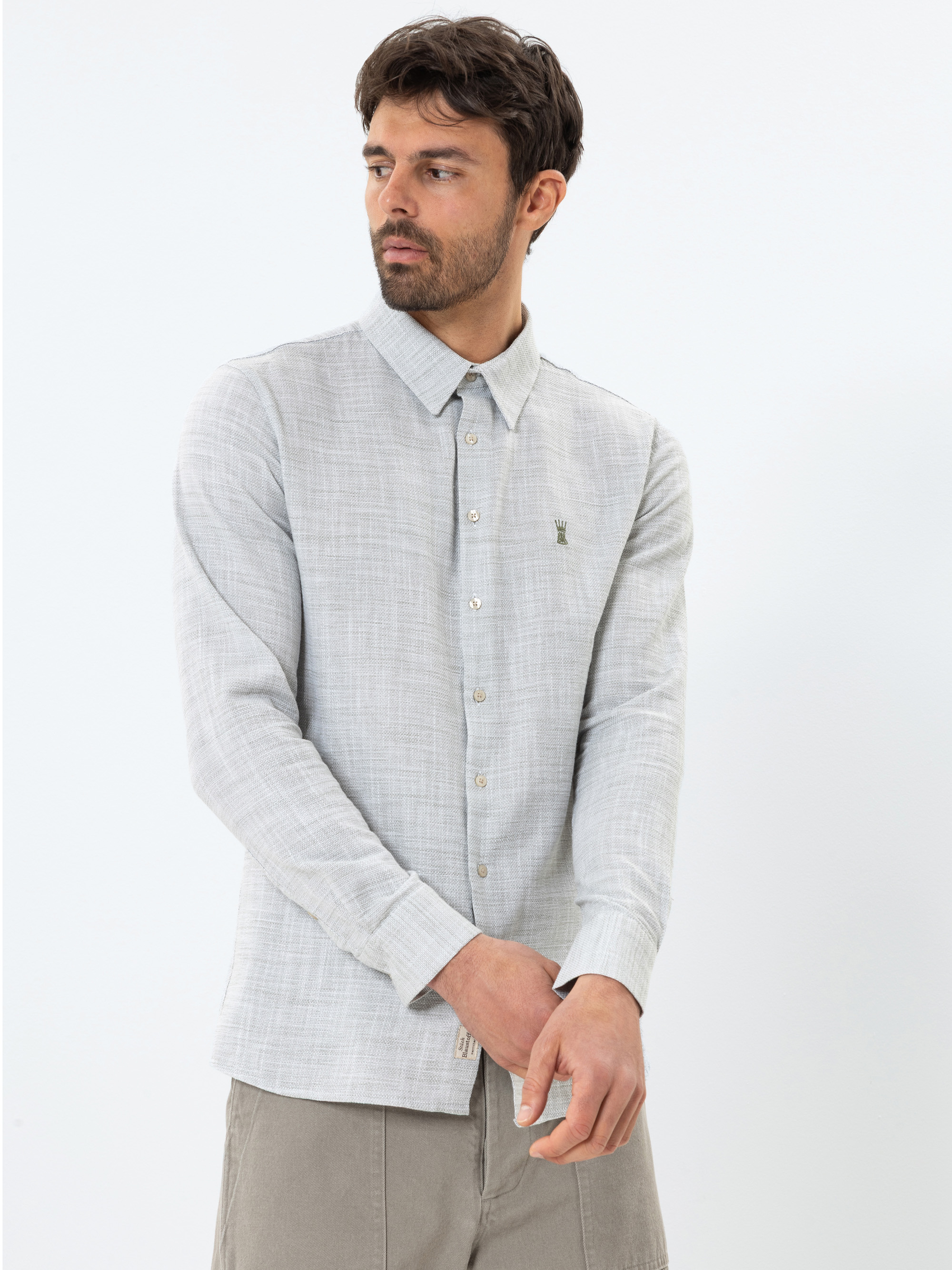 Ein Mann mit kurzen dunklen Haaren und einem Bart trägt ein hellgraues, langärmeliges Button-up-Hemd und eine helle Hose. Er steht vor einem schlichten weißen Hintergrund und schaut leicht nach rechts.