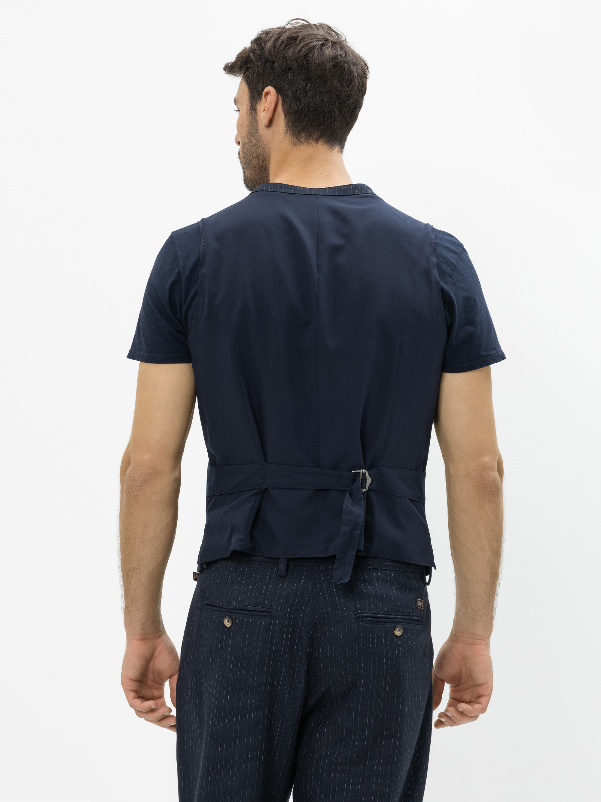 Ein Mann steht mit dem Rücken zur Kamera und trägt ein marineblaues, kurzärmeliges Hemd, eine passende Weste mit Schnalle auf dem Rücken und eine dunkle Nadelstreifenhose vor einem schlichten weißen Hintergrund.