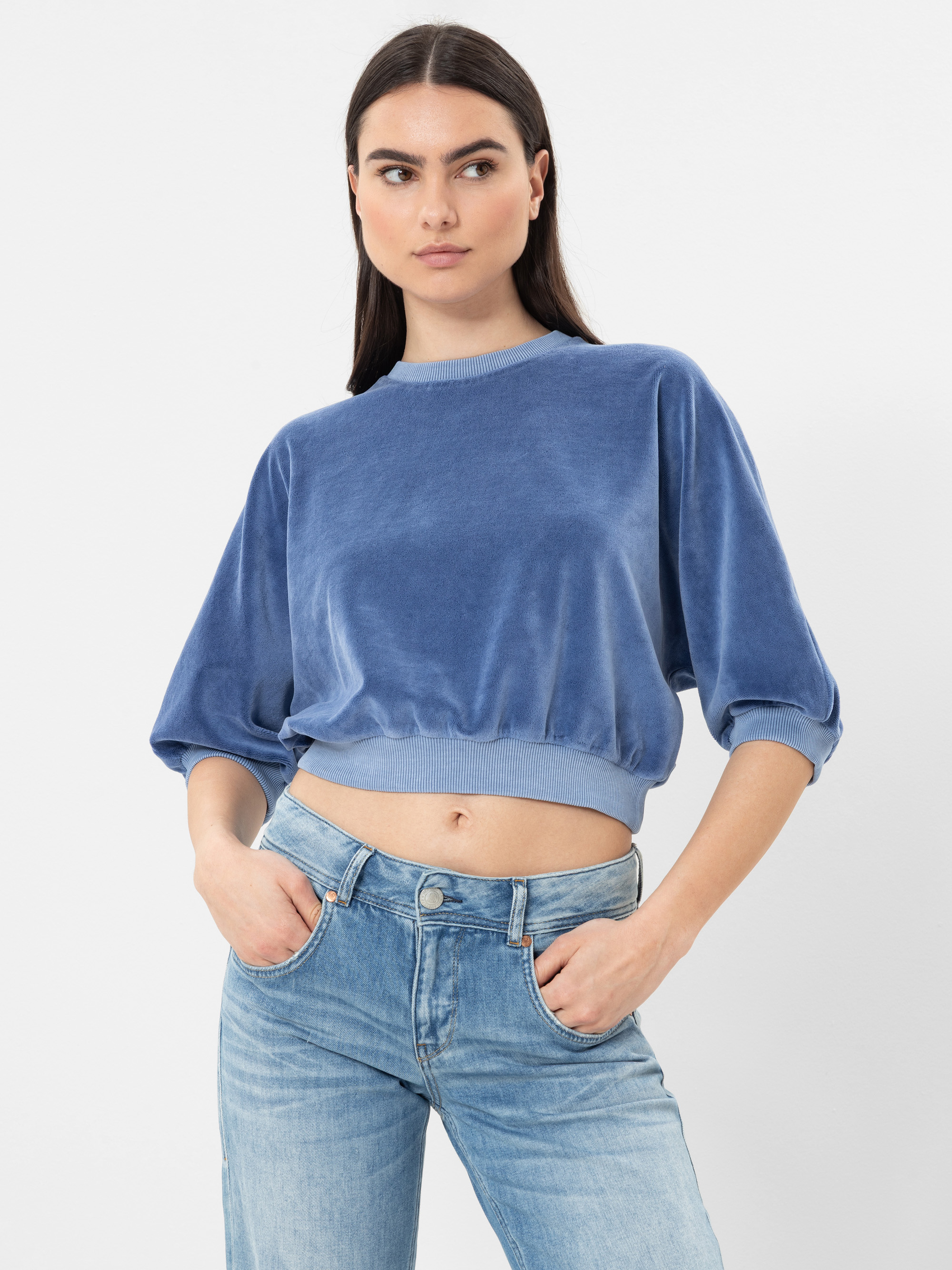 Eine Frau, die ein blaues, samtenes Sweatshirt mit halben Ärmeln und hellblaue Jeans trägt, steht vor einem schlichten weißen Hintergrund und blickt mit neutralem Blick leicht zur Seite.