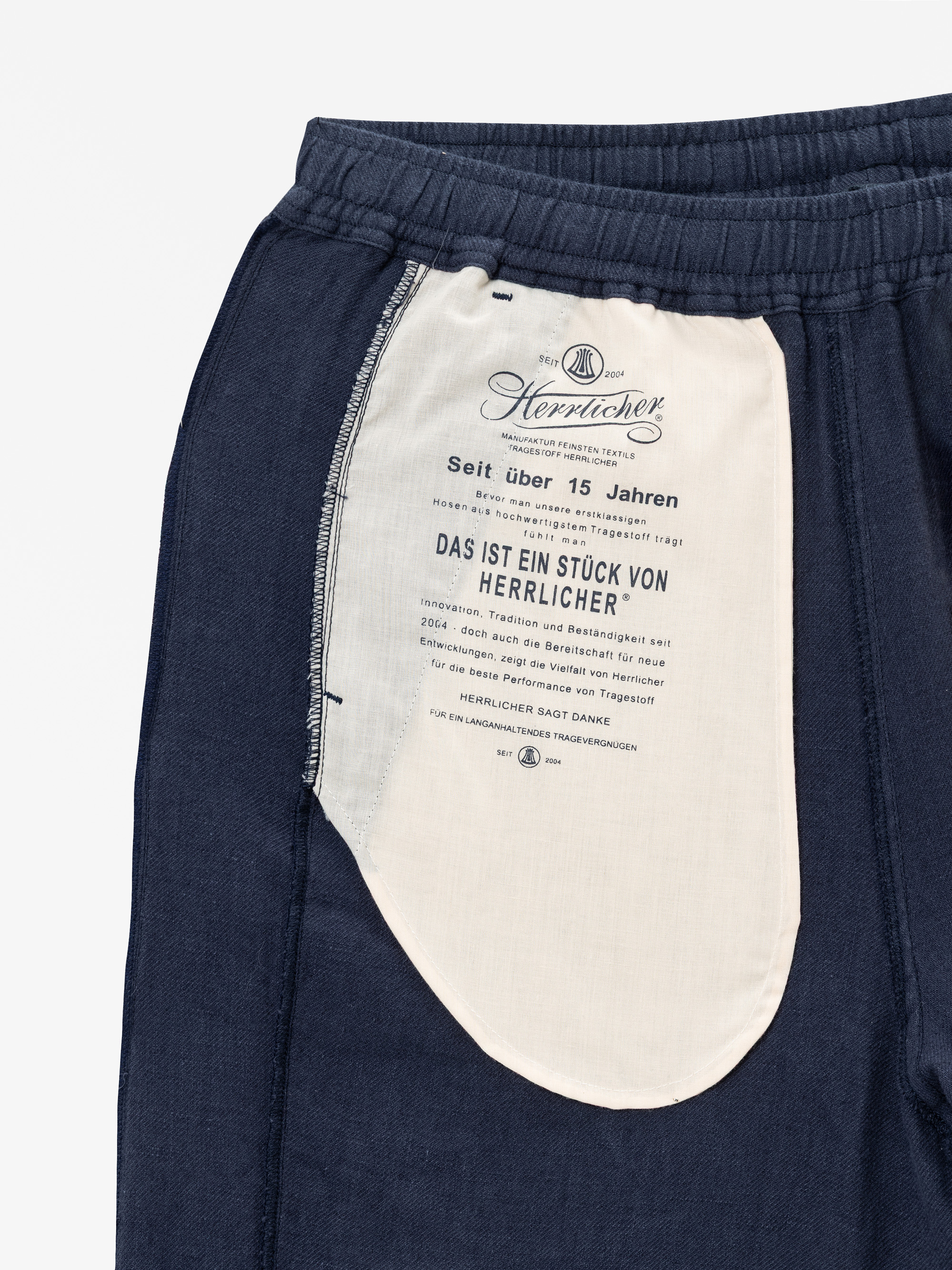 Nahaufnahme der marineblauen Hose, die das Innenfutter der Tasche zeigt, das weiß mit deutschem Text und dem Branding von Herrlicher ist. Der Bund ist elastisch, und die Nähte der Taschen sind auf dem dunklen Stoff sichtbar.