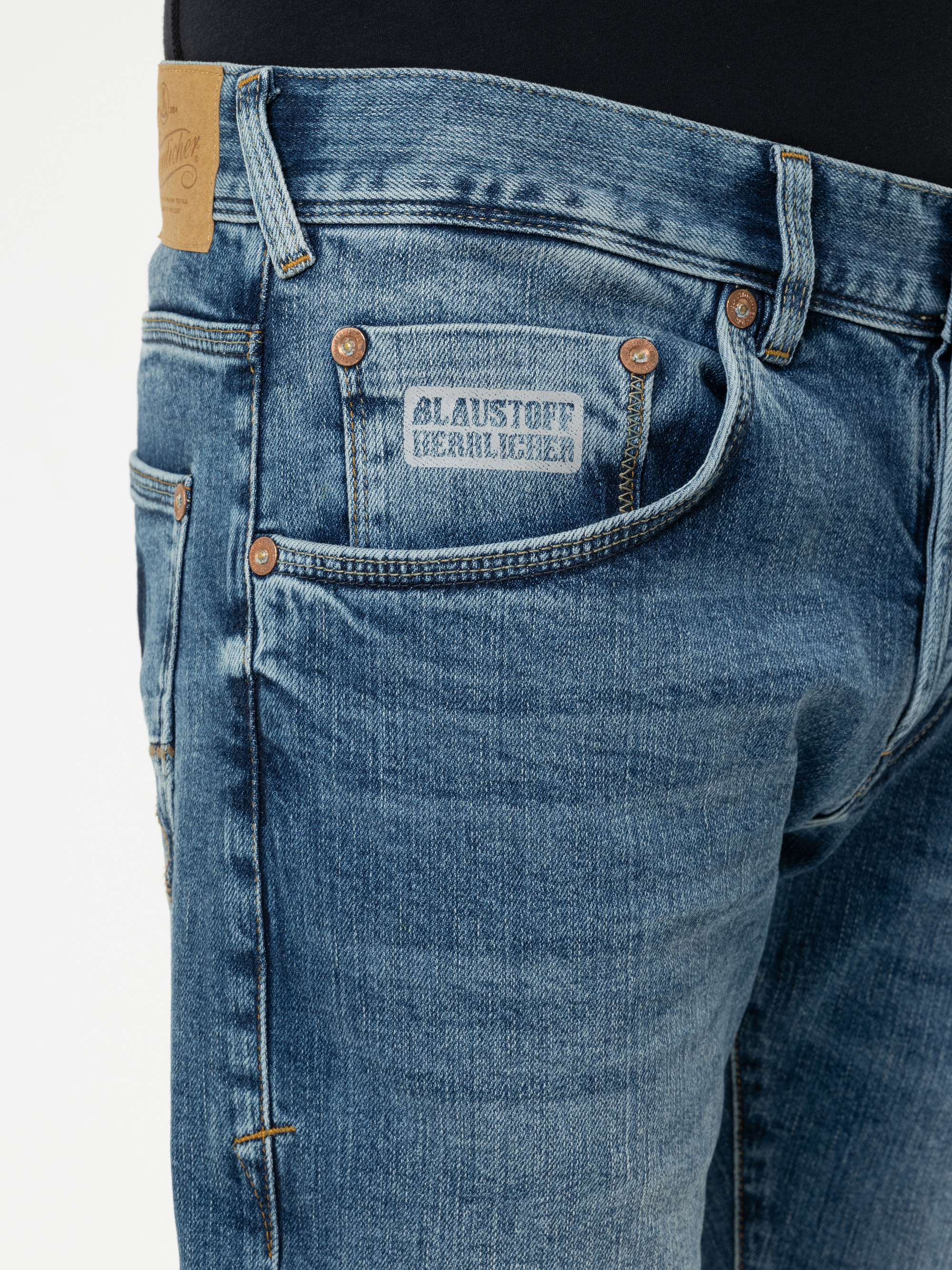 Nahaufnahme einer Person, die eine blaue Jeans mit sichtbaren Nähten, Kupfernieten und einem kleinen Etikett über der Vordertasche mit der Aufschrift AUGUST VERHILLER trägt. Auch ein Teil eines schwarzen Hemdes ist zu sehen.