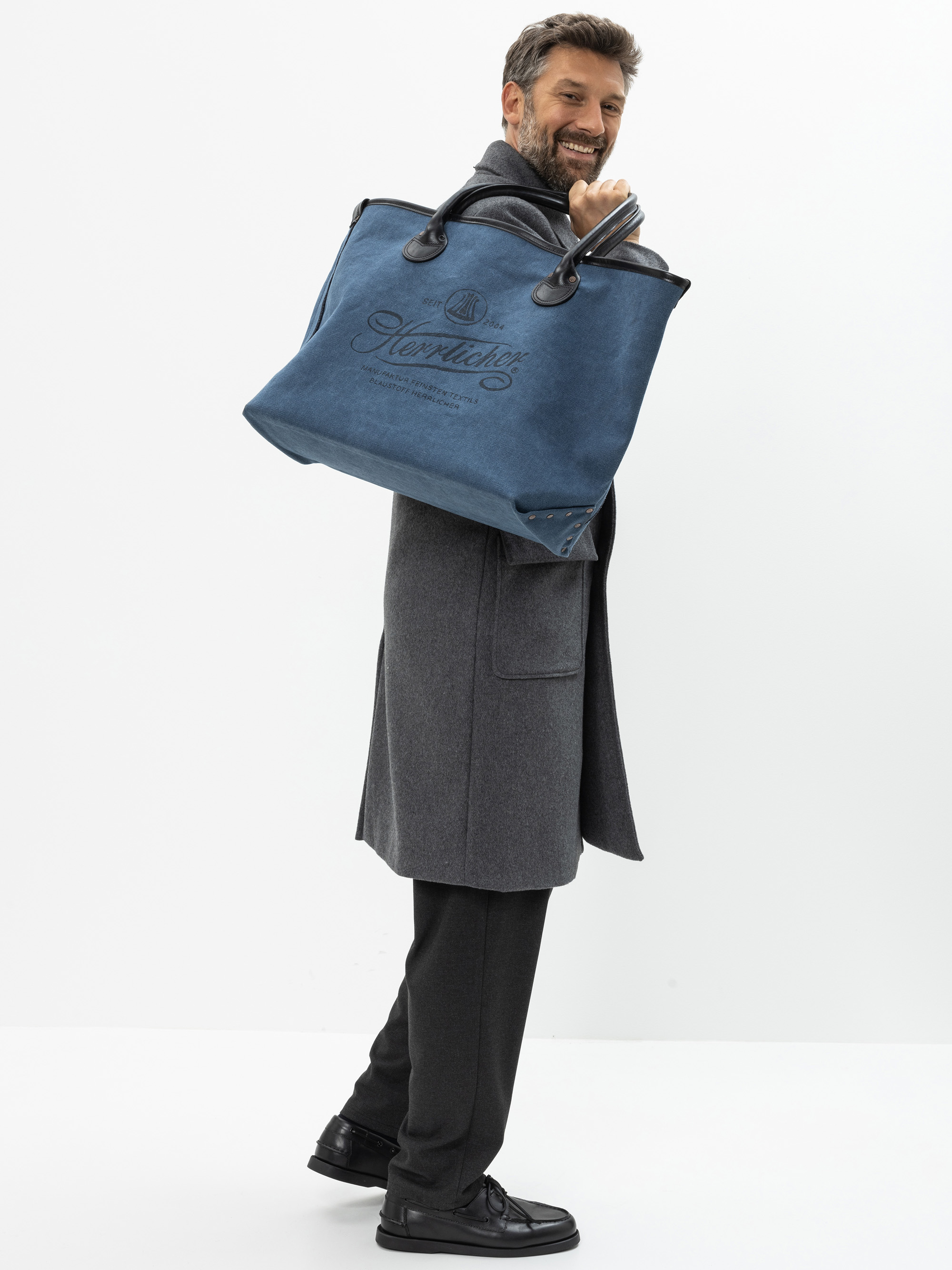 Ein lächelnder Mann in einem grauen Mantel und schwarzen Schuhen hält eine große blaue Tragetasche über der Schulter, während er vor einem schlichten weißen Hintergrund steht.