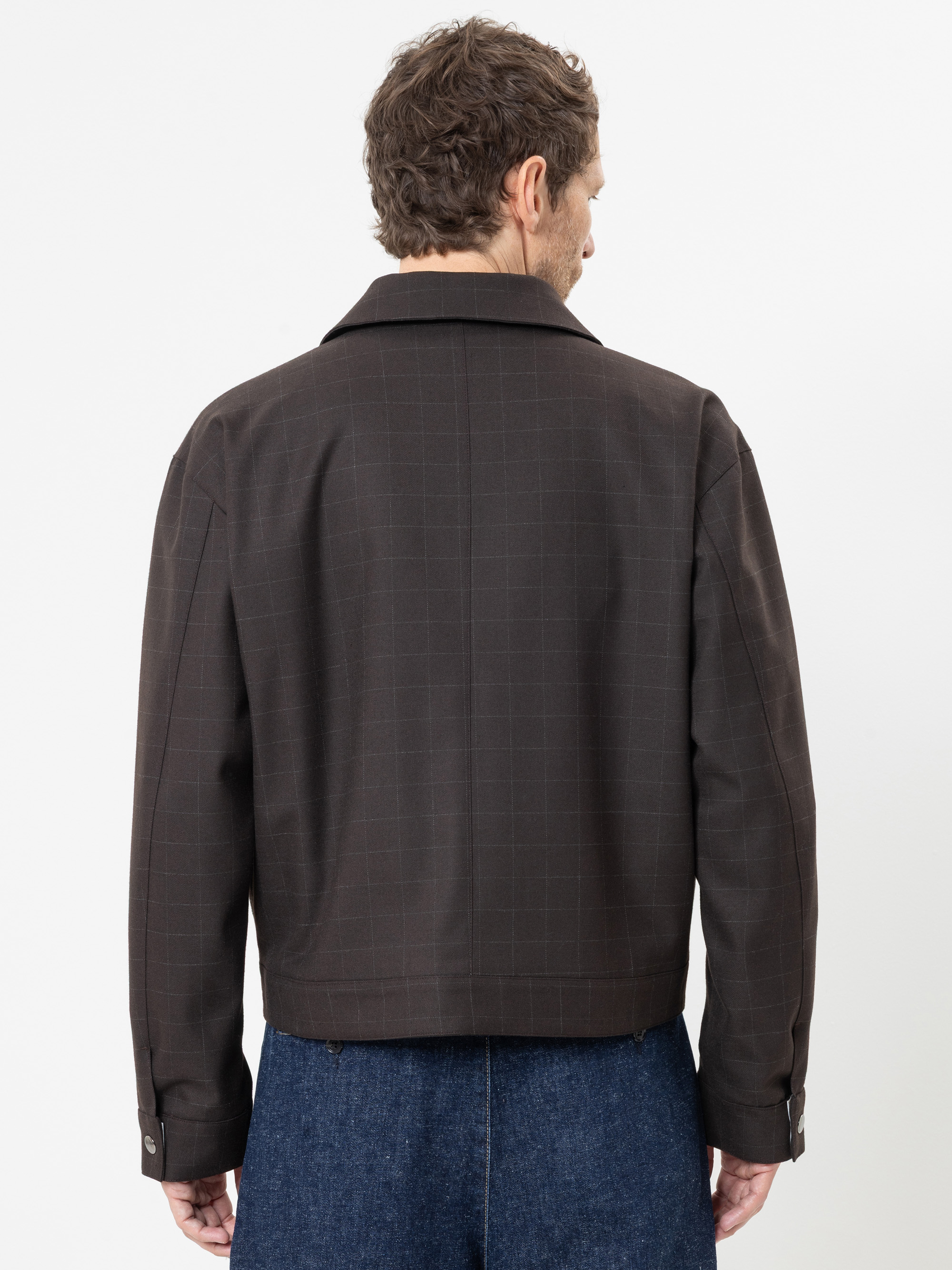 Ein Mann, der mit dem Rücken zur Kamera steht und eine dunkelbraune, langärmelige Jacke mit dezentem Karomuster sowie eine blaue Jeans trägt, vor einem schlichten weißen Hintergrund.