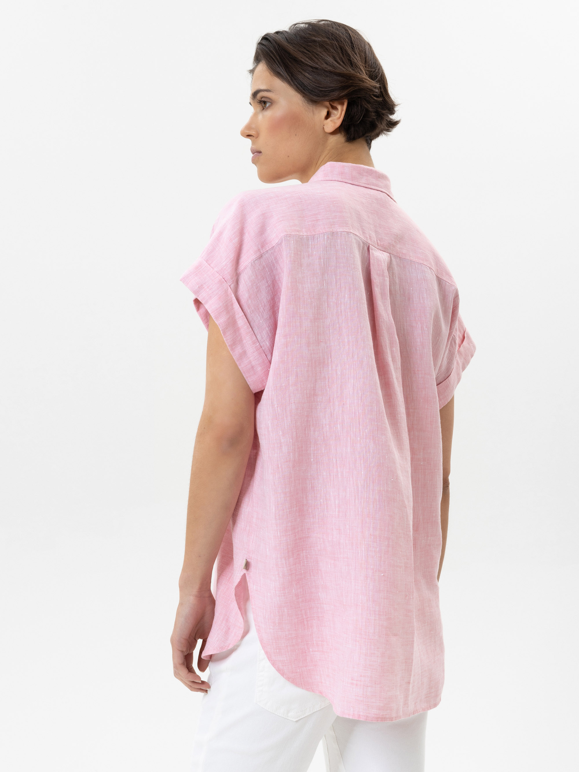 Eine Person mit kurzen braunen Haaren trägt ein locker sitzendes, kurzärmeliges rosafarbenes Hemd und eine weiße Hose. Sie steht mit dem Rücken zugewandt und blickt zur Seite vor einem schlichten weißen Hintergrund.