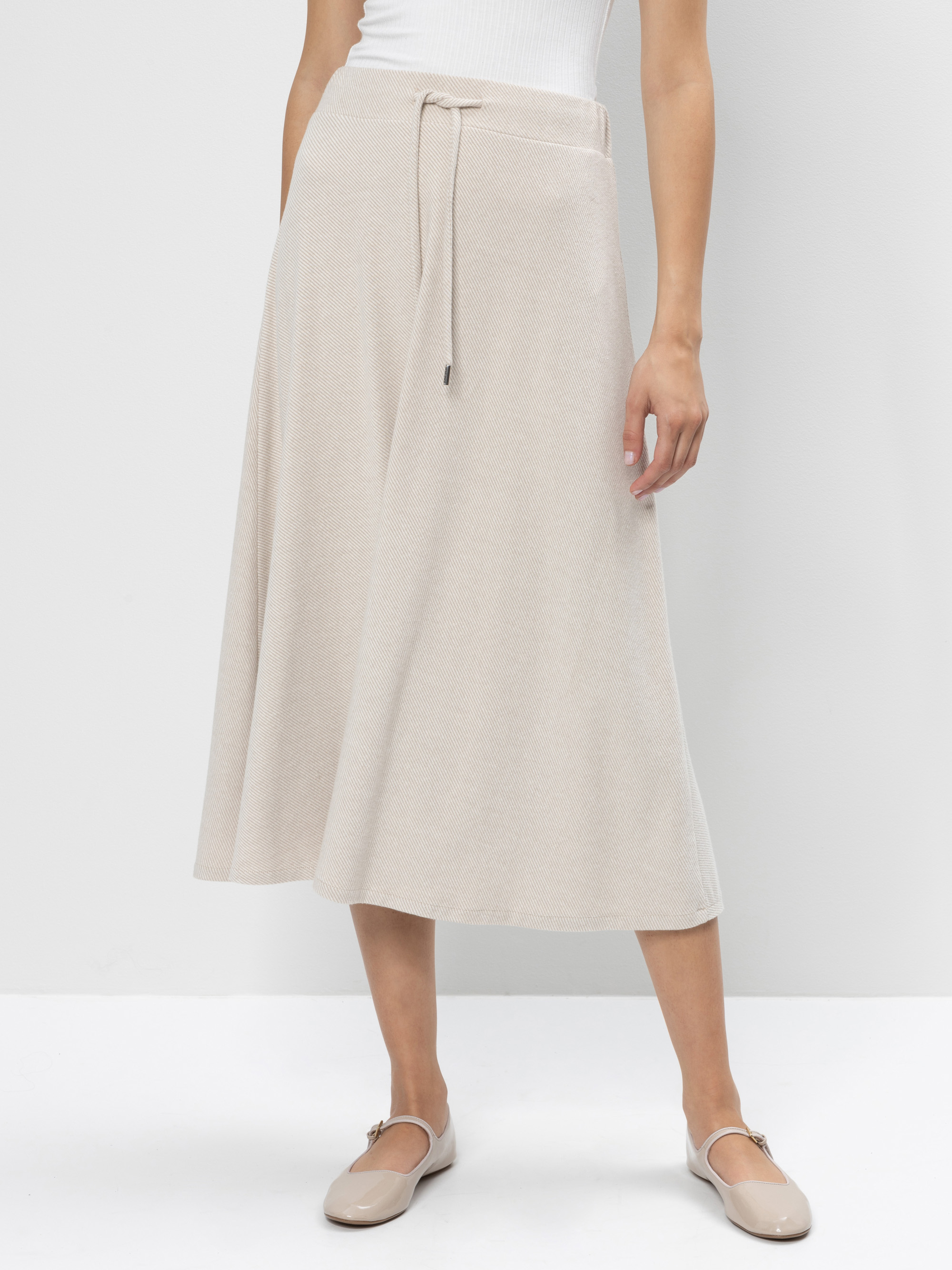 Eine Person, die einen hellbeigen, knöchellangen Rock in A-Linie mit Kordelzug in der Taille, ein weißes ärmelloses Oberteil und beigefarbene Ballerinas trägt, steht vor einem schlichten weißen Hintergrund.